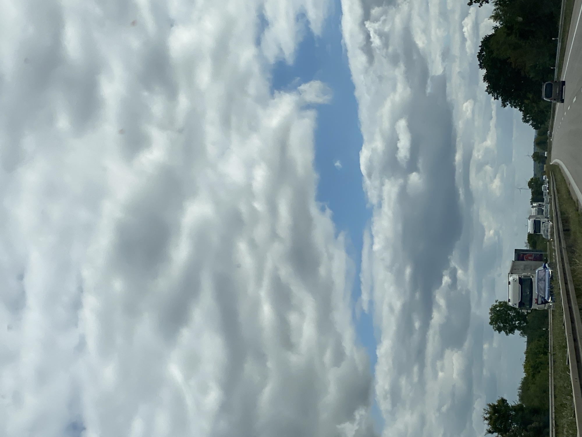 The image depicts a view from a vehicle traveling on a highway in Roßwein, Germany. The sky is filled with a dynamic pattern of grey and white clouds, with patches of blue sky visible. The highway stretches into the distance, with other vehicles, including trucks and cars, present on the road. To the sides of the highway, there are trees and some vegetation, suggesting a rural or suburban setting. The overall impression is one of overcast weather during daytime travel.