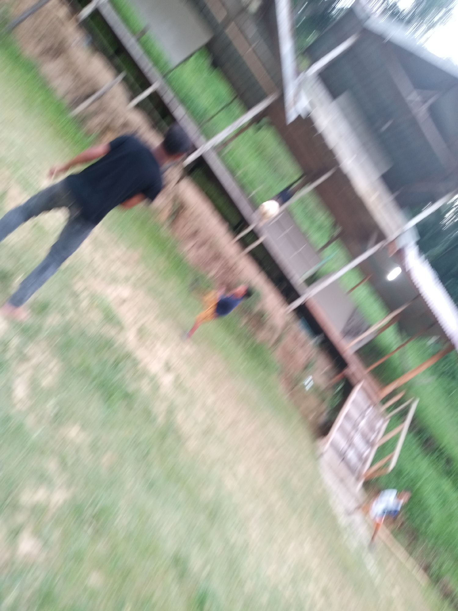 In Lhokseumawe, Indonesia, a blurred image captures an outdoor scene with several individuals and a structure that appears to be a shaded area or a small building with a corrugated roof. The ground is covered in grass and dirt.
In the foreground, a person wearing a dark t-shirt and dark pants is walking. Further into the scene, another person in the background is wearing a light-colored shirt and dark shorts, walking near a wooden staircase. A third person, further in the background, is wearing a blue shirt and possibly shorts or pants. The image's motion blur suggests that the photographer or subjects were in motion. The overall impression is of a casual, possibly rural or semi-rural setting.