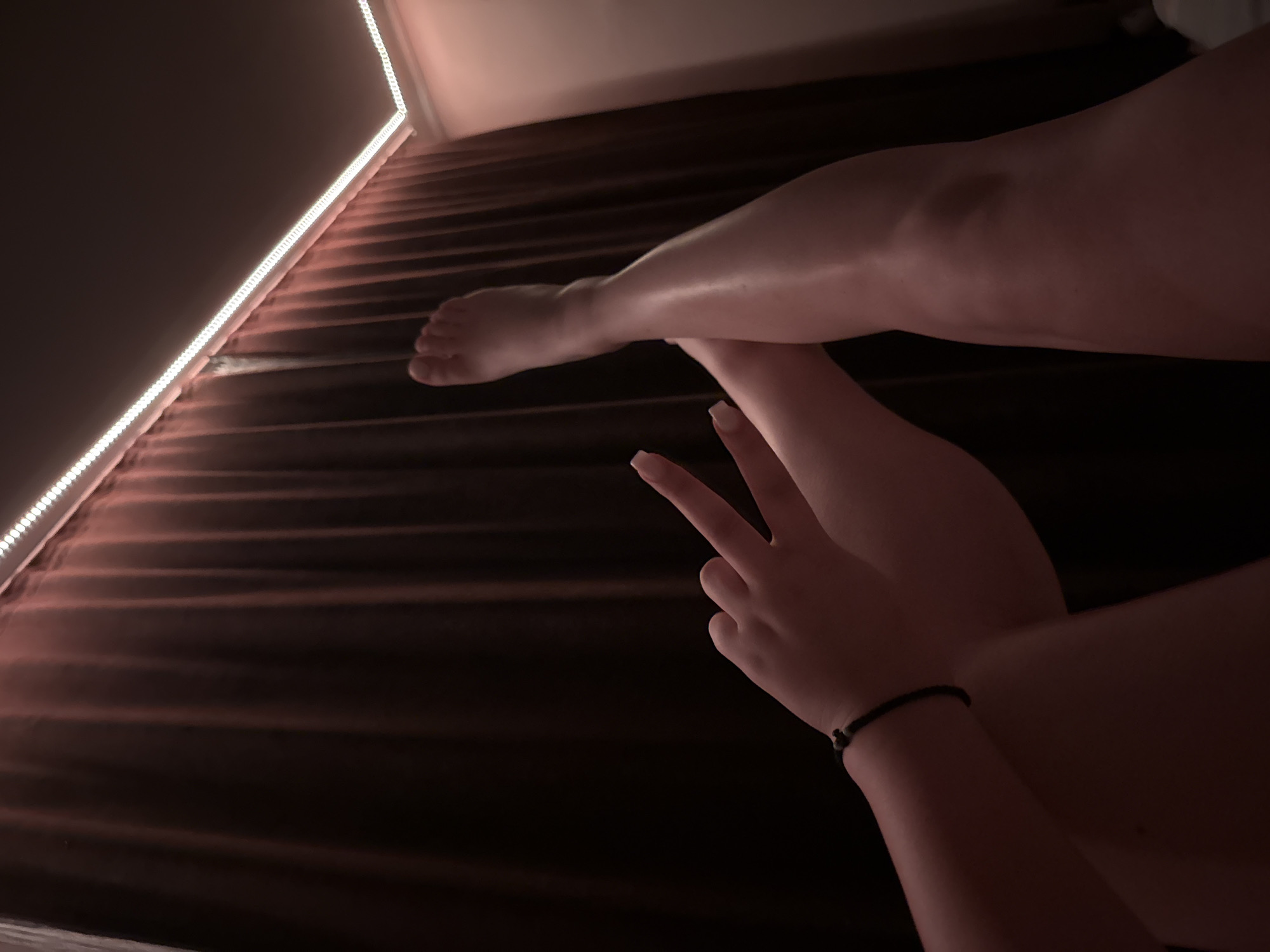 The image captures a close-up, low-angle view of a person's legs and one hand in a dimly lit indoor setting. The person's legs are visible from the thighs down, appearing crossed or overlapping, and are positioned on a textured, dark reddish-brown surface, likely a bedspread with horizontal ridges. One hand is prominently displayed in the foreground, making a peace sign (V-sign) with two fingers extended. A simple, dark cord bracelet is worn on the wrist of this hand.
The scene is illuminated primarily by a strip of bright white LED lights running along the edge of what appears to be a wall or furniture piece on the left side of the frame. This lighting creates strong highlights on the skin of the legs and fingers, contrasting with the deep shadows that envelop much of the background. The overall impression is one of a relaxed, casual moment, possibly taken at night or in a room with minimal ambient light. There is no visible text in the image. The location is Rogosh, Bulgaria.