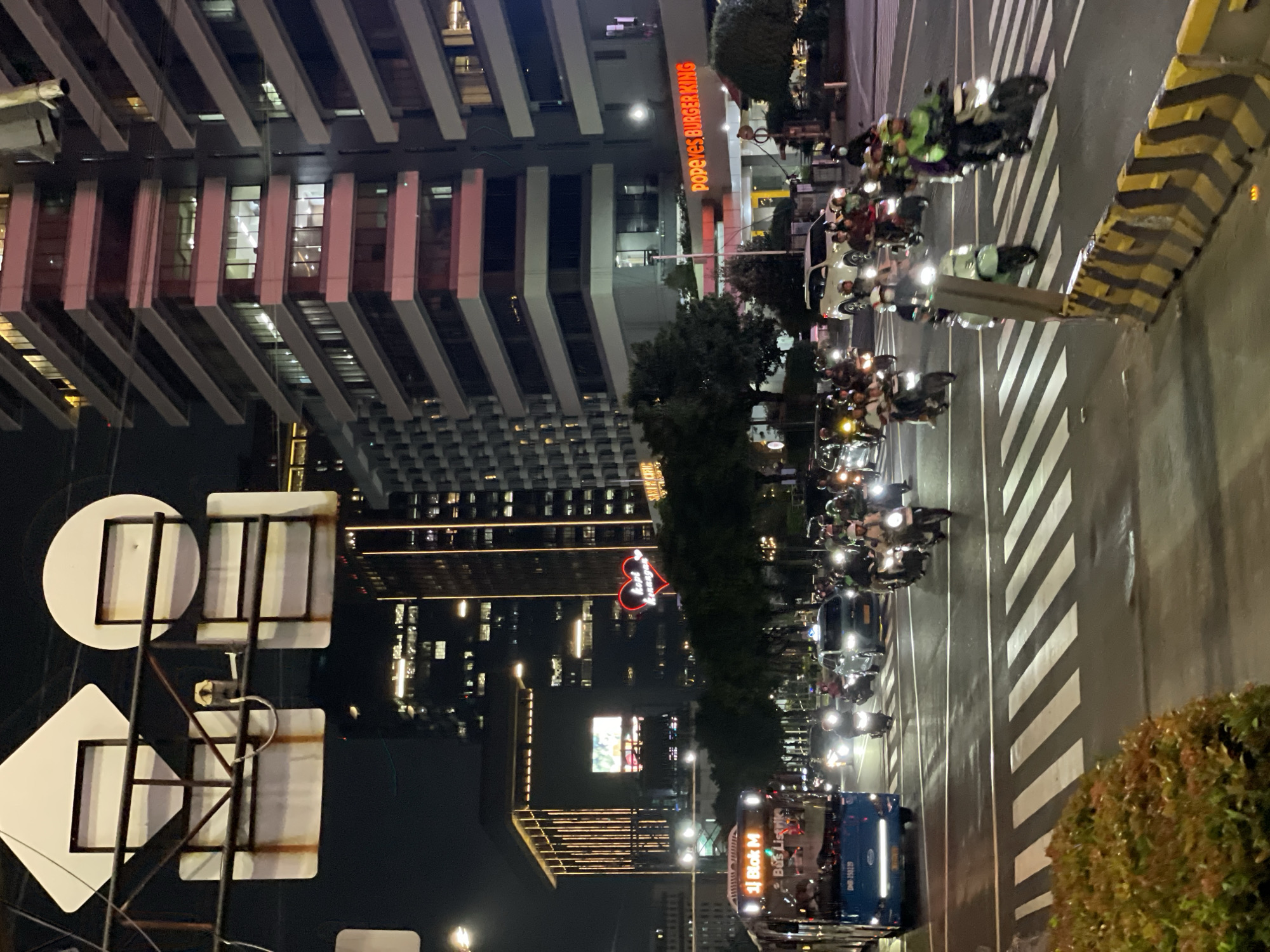 The image depicts an evening street scene in Central Jakarta, Indonesia. A major road is visible, filled with heavy motorcycle traffic, as well as cars and buses. Pedestrian crosswalks are marked with white stripes. Buildings flank the road, some with bright commercial signage (Popeyes Burger King) and illuminated advertisements. A building with a heart-shaped neon sign indicating 'Love' and an illuminated signboard 'Santa Cruz Legacy' can be seen. Street signs and traffic barriers are visible. The overall atmosphere suggests a bustling urban environment at night.