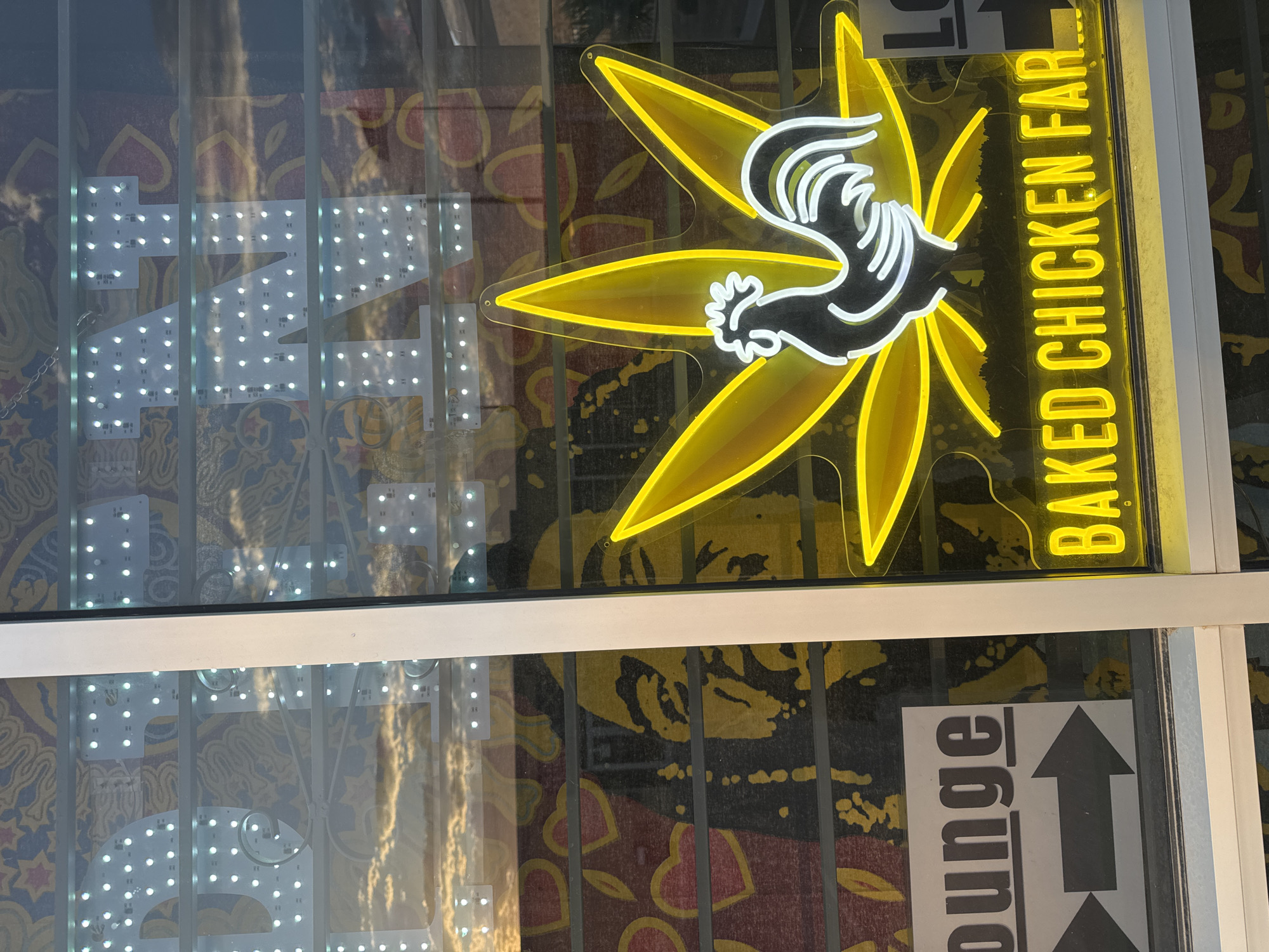 The image shows the exterior of a business in Sunland Park, New Mexico, USA. A neon sign in yellow and white depicts a cannabis leaf with a rooster silhouette in the center, and the text "BAKED CHICKEN FAR..." below it. Behind the neon sign, a large graphic of a face, possibly of Bob Marley, is visible. To the left, a sign reads "lounge" with an arrow pointing to the right. The business appears to be located on a street, as indicated by the reflection of the sky in the window. The overall impression is that of a quirky, potentially cannabis-themed establishment.