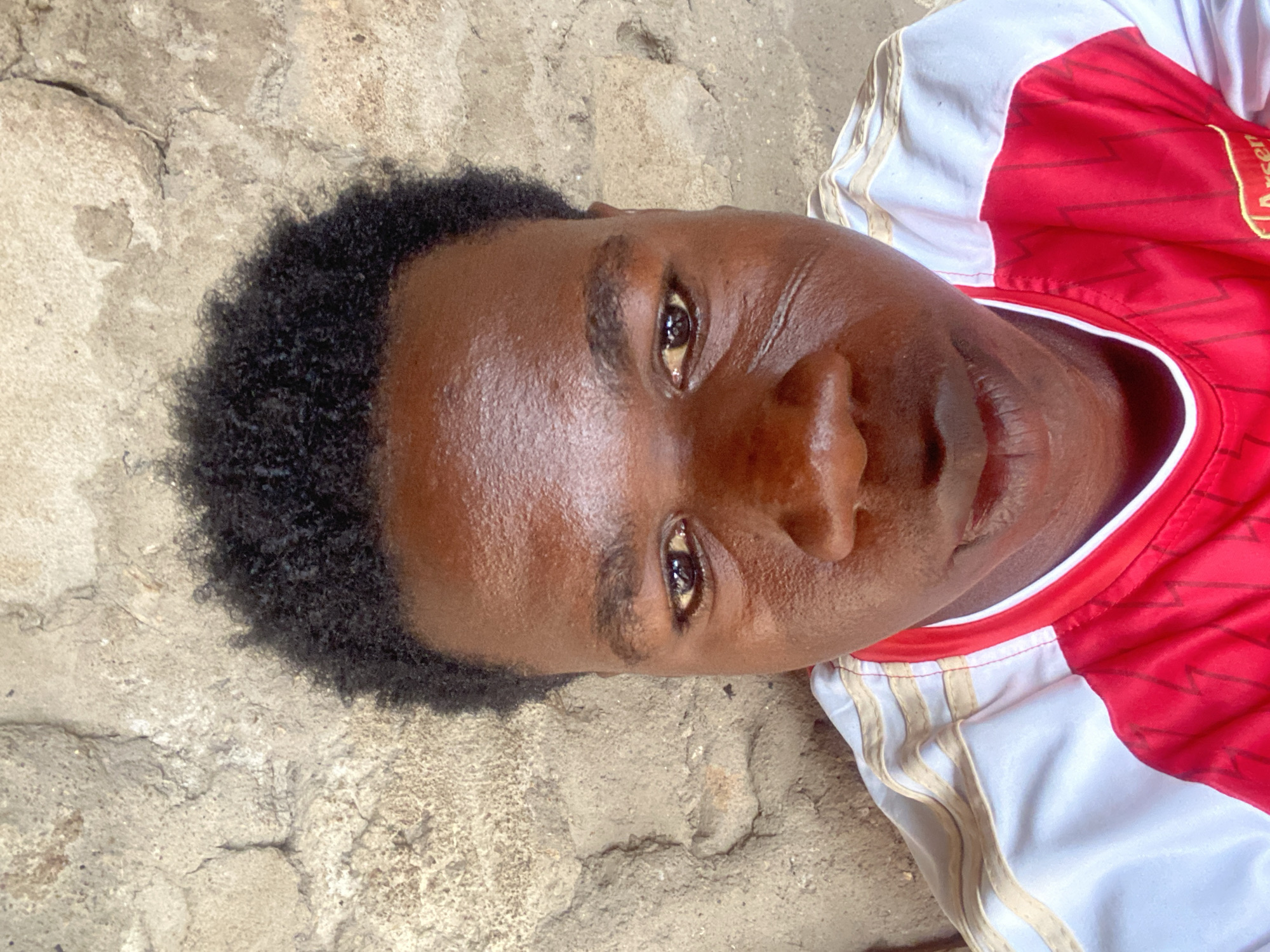 A close-up, angled shot depicts a young, dark-skinned male lying on a textured, concrete surface. He has short, dark hair and is wearing a red and white soccer jersey. The jersey has vertical stripes. The location is presumed to be in the city of Mararaba, Nigeria.