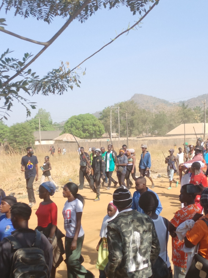 The scene depicts a daytime outdoor gathering or procession on a wide, unpaved dirt path in Kpak, Nigeria. The environment is characterized by dry, reddish-brown ground, sparse yellowed grass, and scattered trees, with prominent hills visible in the distant background under a clear, hazy blue sky. Several single-story buildings, including one with a round, conical roof, and utility poles are present in the mid-ground.
Approximately 50 to 70 individuals, predominantly young adults of varying genders, with some children, are visible. They are casually dressed in t-shirts, long-sleeved shirts, trousers, and shorts of various colors. A significant number of individuals, particularly in the mid-ground, are carrying long, slender wooden sticks or staves, held over shoulders or in hands.
Key individuals include:
* In the mid-ground left, a male wearing a black t-shirt, dark trousers, and a black face mask walks.
* Among the group carrying sticks, one male wears a black uniform-like top with yellow accents and a dark cap.
* In the foreground right, a person wearing a camouflage jacket and a striped white beanie faces away, with another individual in an orange patterned shirt holding a smartphone, seemingly recording.
* In the foreground left, individuals include a female in a red t-shirt, a female in a white t-shirt with "100" printed on it, and a male with a backpack.
The individuals appear to be walking or standing, generally moving or oriented towards the right side of the frame, indicating a collective movement or gathering.