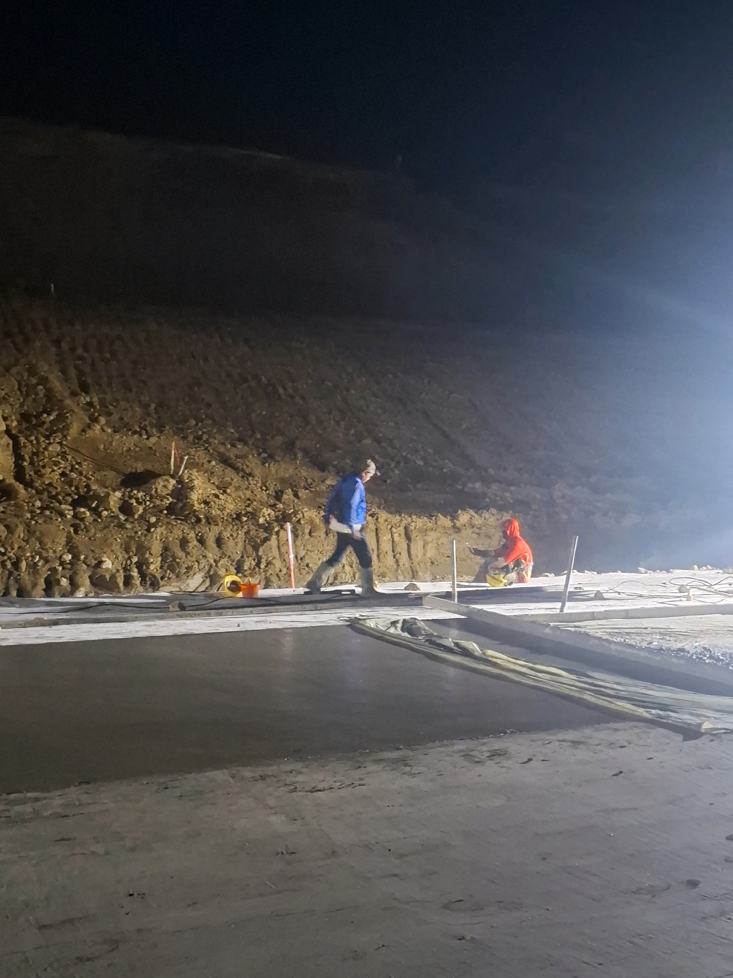 This image captures a nighttime scene at a construction site in the city of Karangtengah, Indonesia. Two individuals are present, engaged in what appears to be concrete work. The person on the left is walking away from the camera, wearing a blue jacket and dark pants, with a white bag slung over their shoulder and dark boots. The person on the right is kneeling, wearing a bright orange hooded jacket and dark pants, and is actively working with a tool or material on the ground.
The environment is characterized by a steep, rocky embankment on the left side of the frame, illuminated by artificial light. In the foreground, a freshly poured concrete surface extends horizontally, bordered by wooden planks. Several orange and yellow buckets, along with orange-tipped stakes, are visible near the embankment, suggesting ongoing construction or surveying activities. The overall lighting suggests that work is being carried out at night, with a strong light source casting a bright glare on the right side of the image and illuminating the workers and the surrounding area. The ground is a mix of concrete and dirt, with some visible debris and footprints.