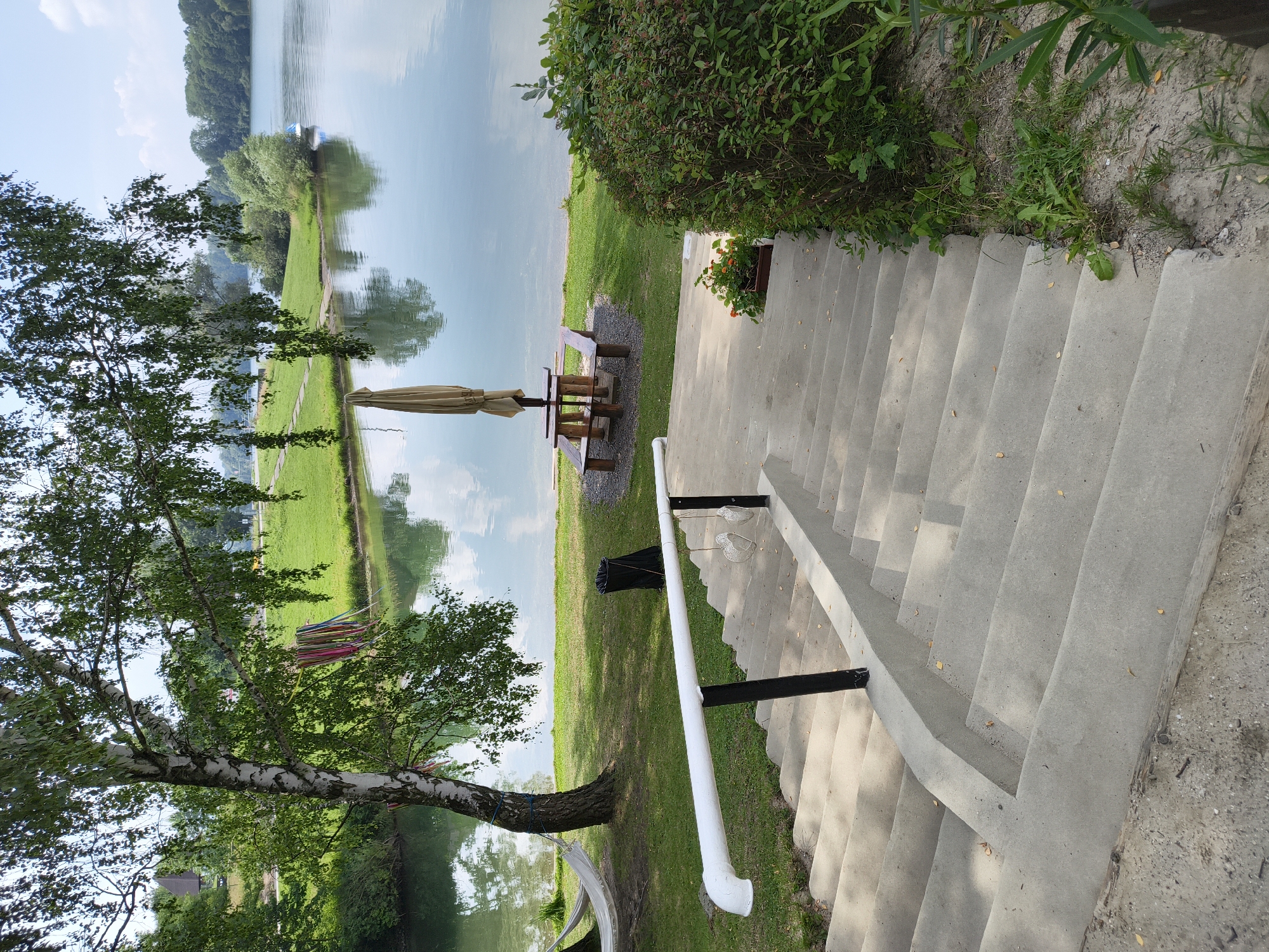 The image shows a concrete staircase leading down to a grassy area next to a calm body of water, likely a lake or river. On the grass, there is a picnic table with an umbrella, a black trash can, and a white hammock strung between two trees. One of the trees has colorful ribbons tied to its branches. In the water, there is a small white boat. The scene appears to be a recreational area on a sunny day with some clouds in the sky. The stairs are in the foreground and take up a significant portion of the image. Lush green trees and foliage surround the water and the grassy bank.