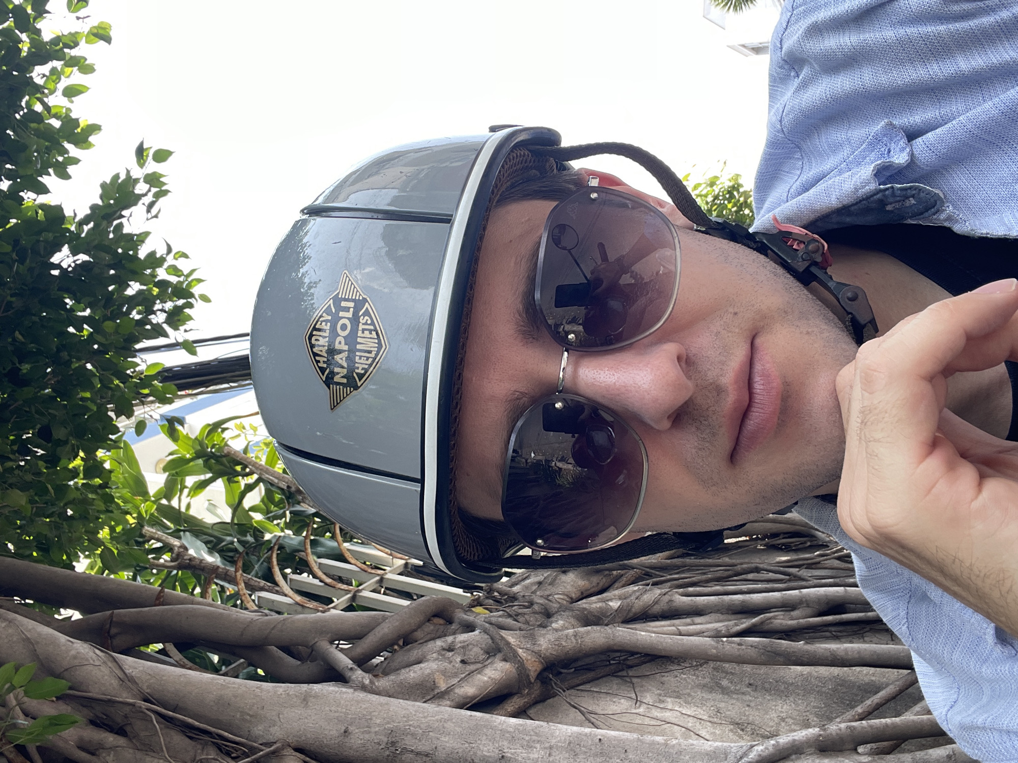 A man wearing sunglasses and a Harley Napoli helmet is pictured outdoors. The surrounding environment includes tree roots in the foreground, with green foliage and hints of urban infrastructure (power lines) in the background, consistent with the city of Nha Trang, Vietnam. He is wearing a light blue shirt. The man's face is partially visible, and his hand is near his face.
