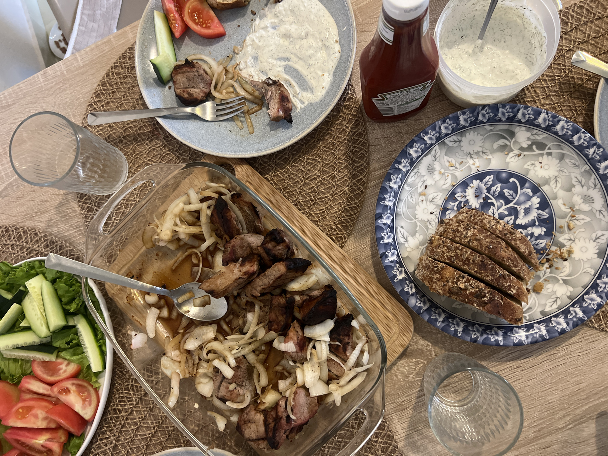 The image shows a table with a meal. There is a clear glass baking dish filled with cooked meat and onions, a plate with sliced brown bread, a plate with sliced tomatoes, cucumbers, meat, and a white sauce, a bottle of ketchup, a container of white sauce with a spoon, and two empty glasses. The table is covered with a patterned mat. The scene appears to be indoors, possibly in a home. There is no indication of the location being in Bar, Montenegro. There are no people visible in the image.