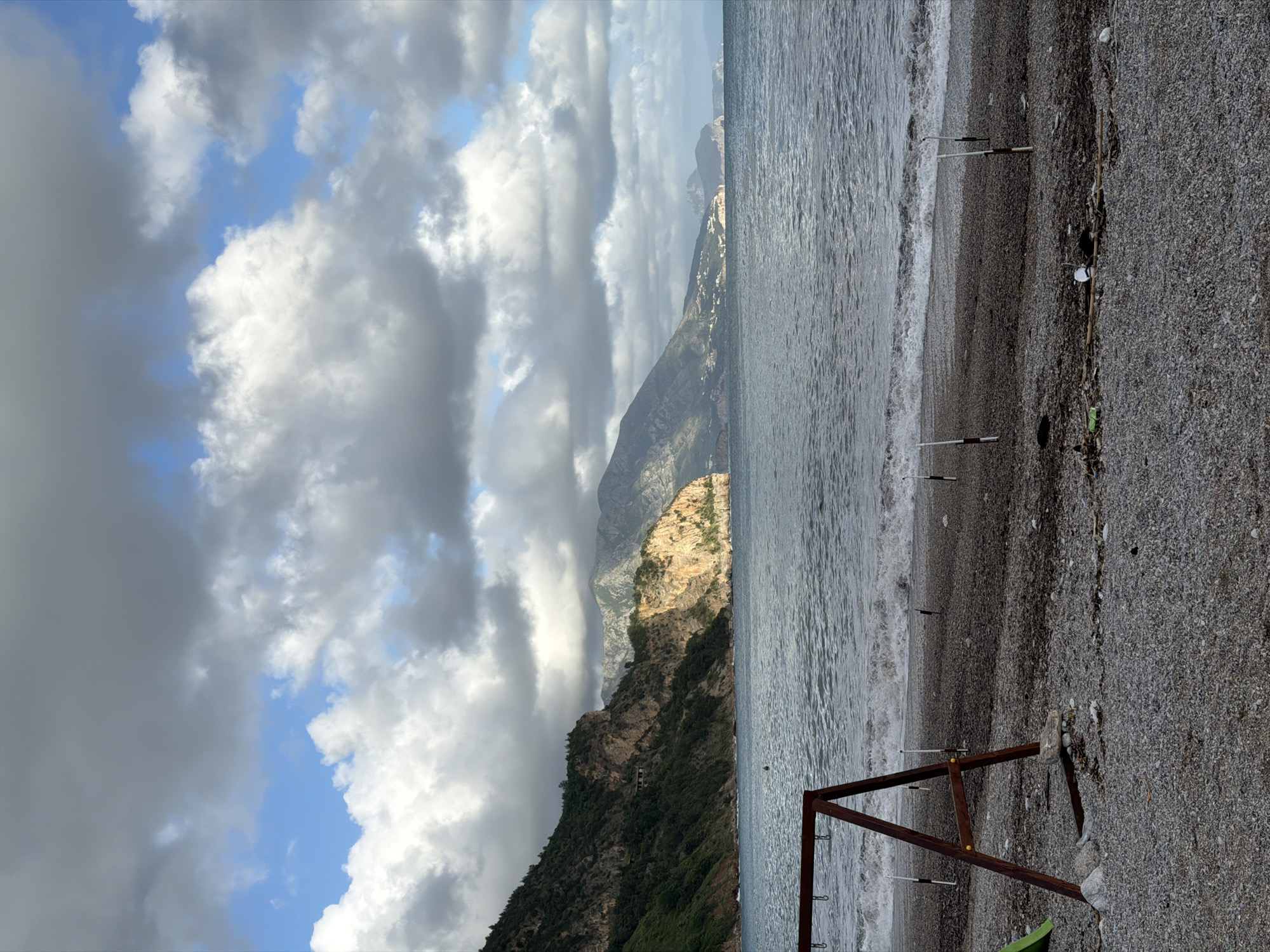 The image depicts a beach scene in or near Prijevor, Montenegro. The sky is filled with dramatic clouds, transitioning from dark gray to white with patches of blue sky visible. In the background, a mountain range rises from the sea. The slopes are covered with vegetation. The sea is choppy. The beach is composed of gray pebbles. In the foreground, a metal structure, possibly part of a beach facility or equipment, stands on the shore. Fishing poles are installed.