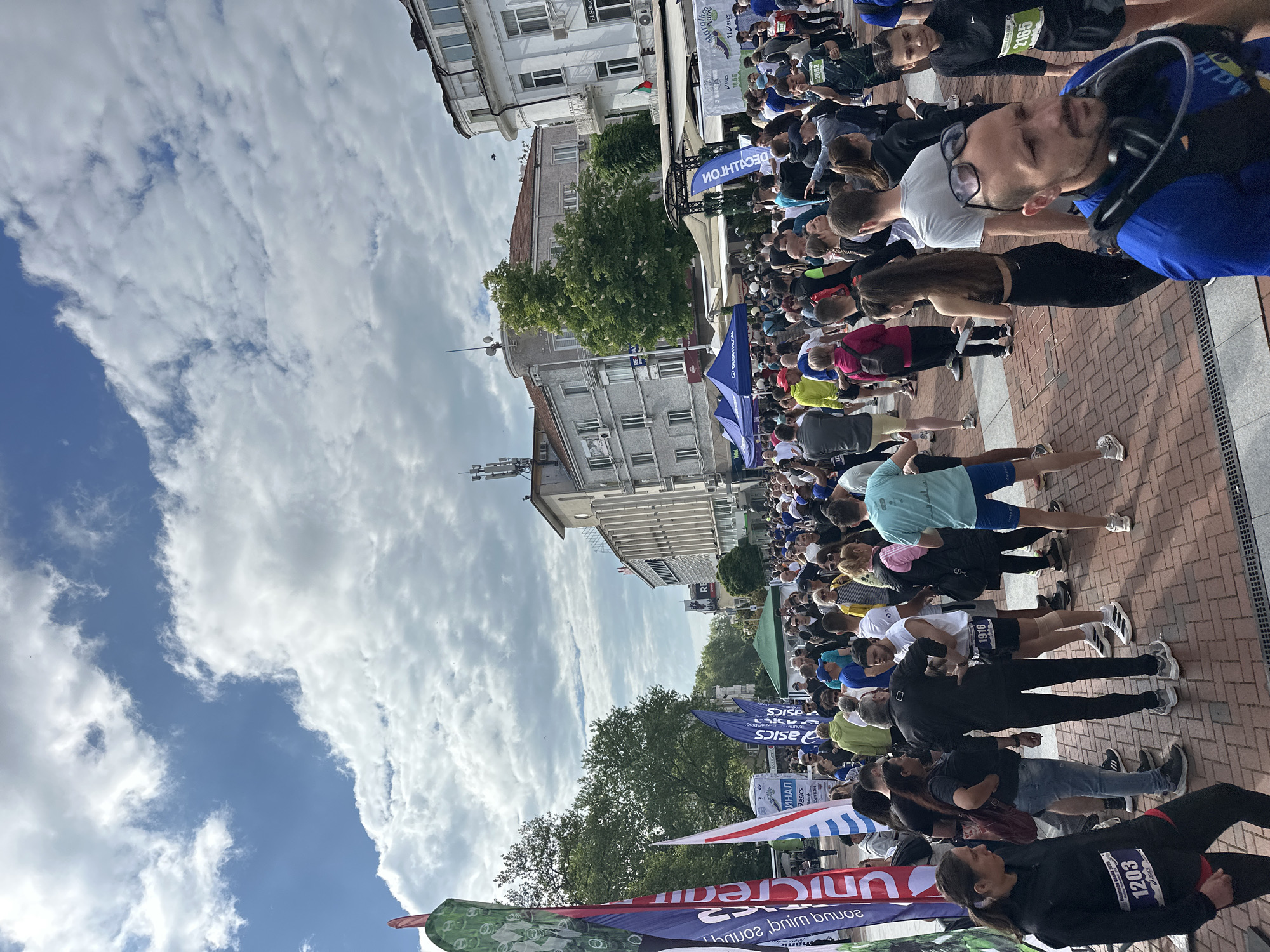 The image depicts a large group of people gathered on what appears to be a pedestrian street in Varna, Bulgaria. The crowd is likely participants in a marathon, as evidenced by the presence of runners with numbered bibs, event banners, and brand logos such as "ASICS" and "UNICREAM". Buildings line one side of the street, suggesting an urban setting. The sky above is filled with clouds, indicating the weather may be fair. Several event tents are visible. The participants are a mix of male and female. The banners appear to be written in Bulgarian.