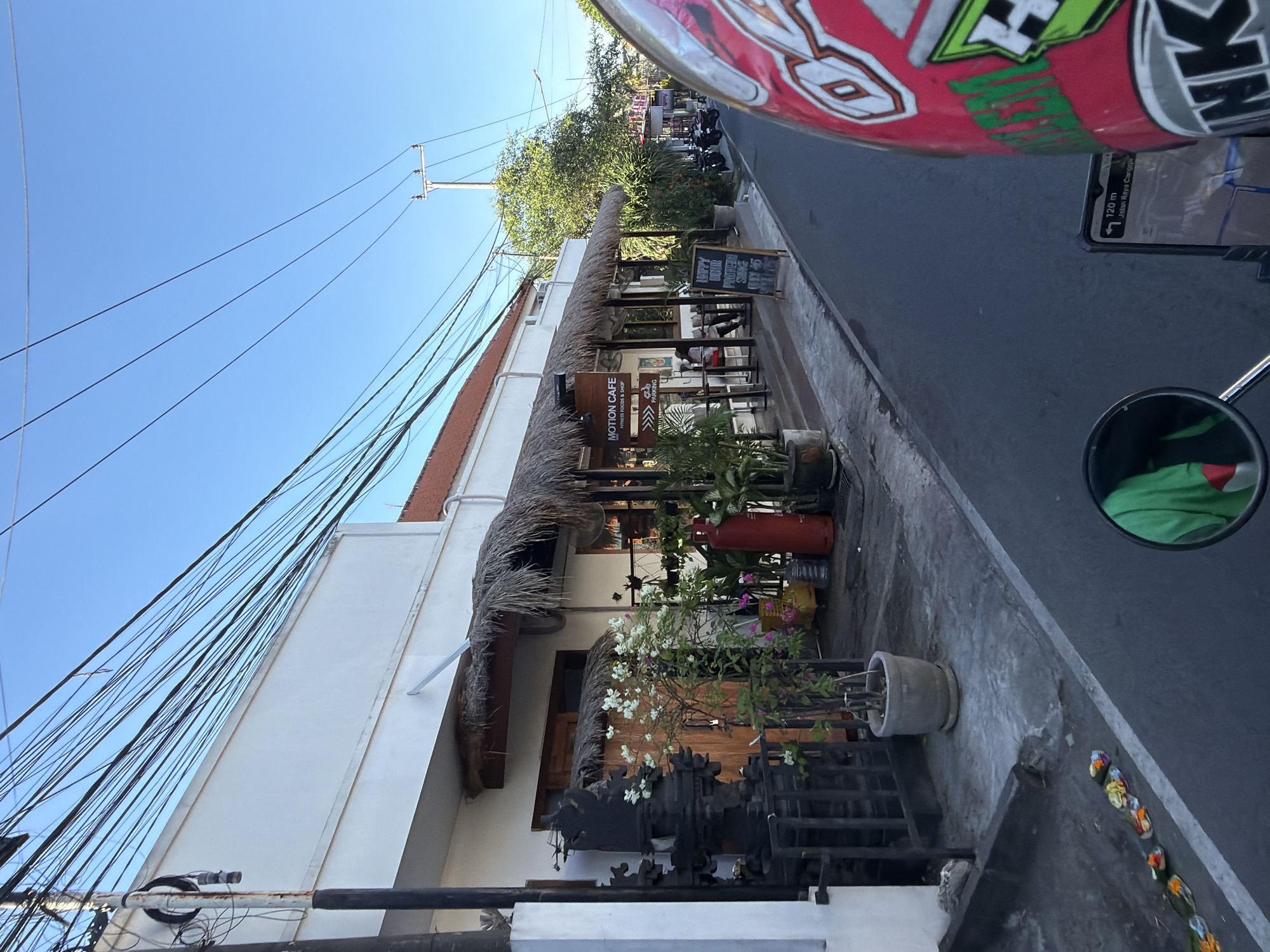 Here is a detailed description of the image:
The image captures an outdoor view of "Motion Cafe", a fitness food and shop in Mengwi, Indonesia. The cafe's storefront is adorned with a thatched roof that extends over an outdoor seating area. A black sign with white letters reads "MOTION CAFE, FITNESS FOODS & SHOP" and a smaller sign indicates parking is available.
The building has a clean white exterior, and numerous electrical wires crisscross the sky above. Potted plants line the pathway and create a natural barrier to the street. A street sign displaying "Jalan Raya Canggu" is visible, confirming the location.
A helmet, partially visible in the upper right of the image, suggests the photo was taken from a moving vehicle, possibly a scooter or motorcycle.
