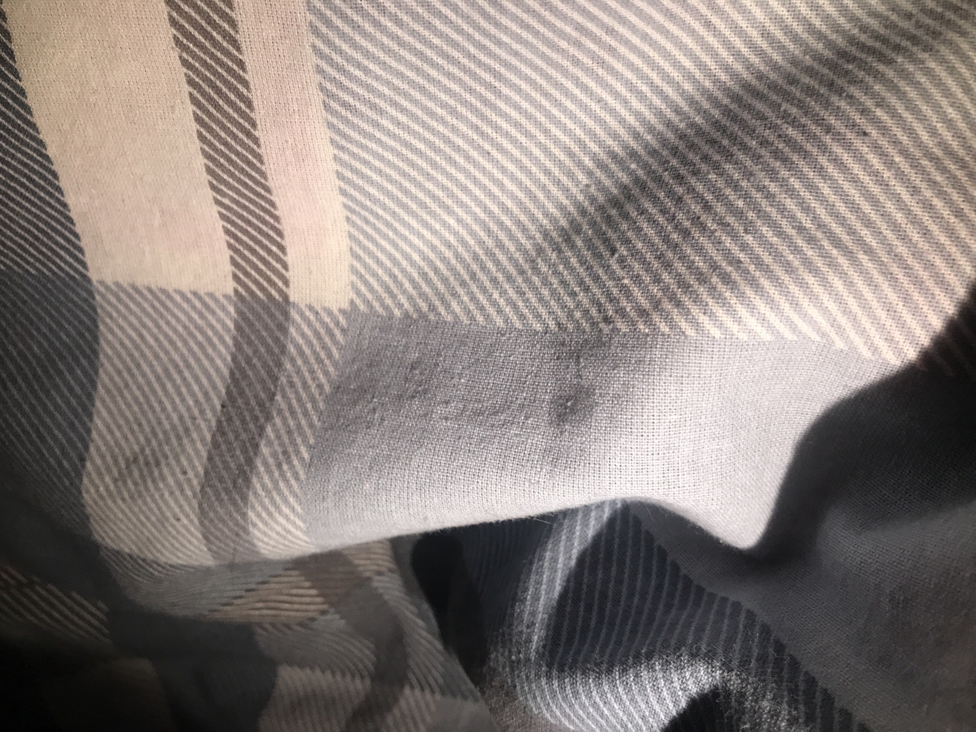 The image shows a close-up of a folded piece of fabric. The fabric has a plaid pattern consisting of white, light gray, and dark gray stripes forming squares and rectangles. The fabric is folded in a way that creates shadows and obscures some of the pattern. There are no people visible, and no information about the location can be inferred from the image.
