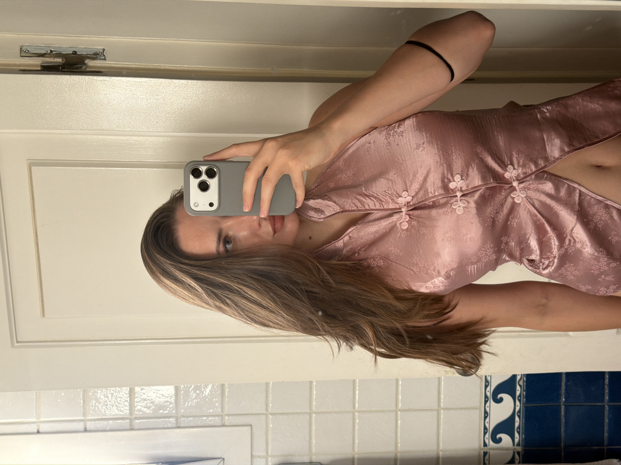 The image shows a person holding a smartphone and taking a selfie in what appears to be a bathroom. The person has long, brown hair and is wearing a pink, satin halter top with a floral pattern and traditional Chinese knot buttons. The background shows white tiled walls and a white door with visible hinges. There is no indication that this location is in Sharm El Sheikh, Egypt.