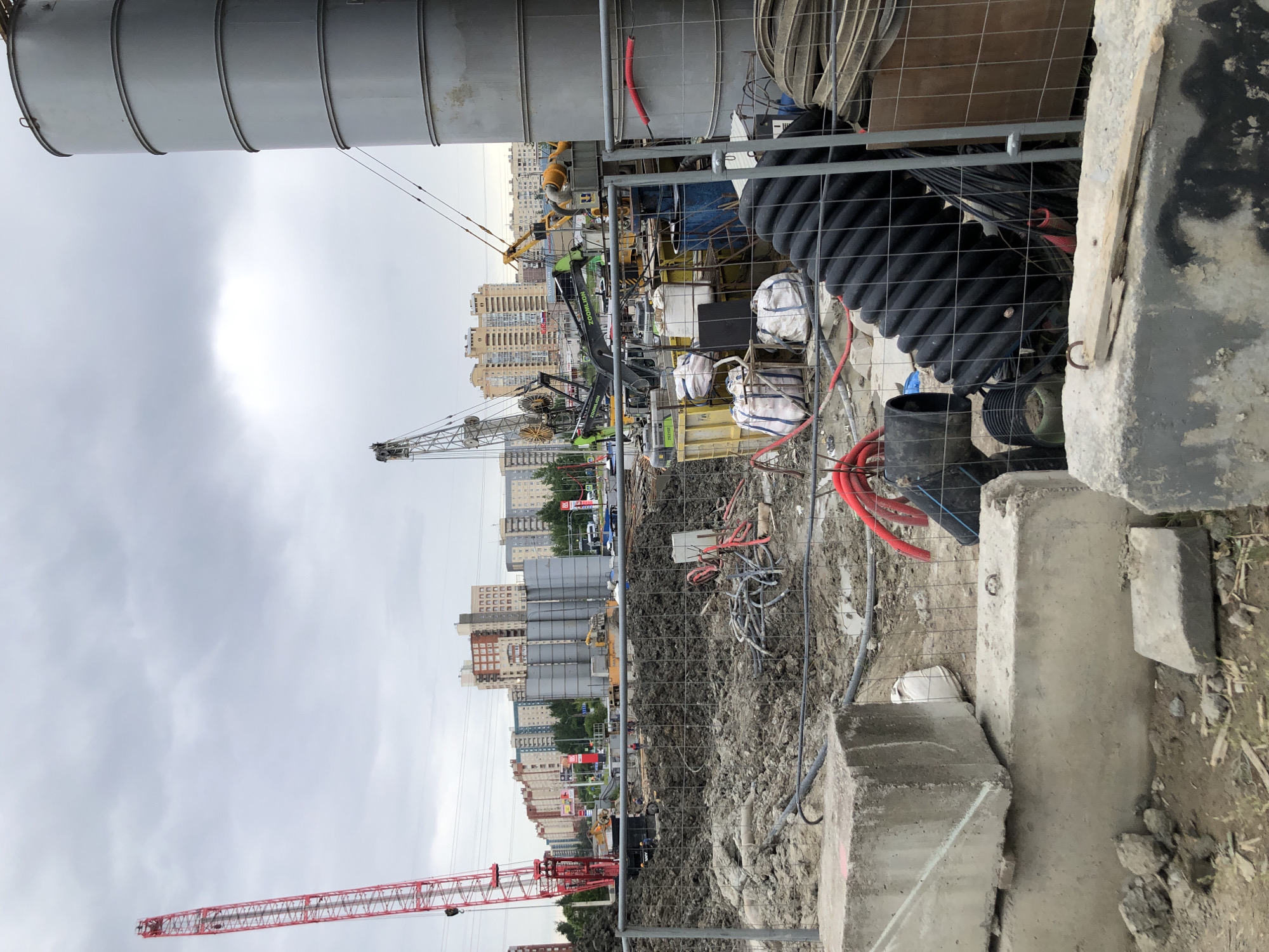 This is a photograph of a construction site in Saint Petersburg, Russia, under a cloudy sky. The foreground features several concrete blocks, some broken, and a section of chain-link fence. Behind the fence, a large pile of dark, wet dirt dominates the mid-ground, with various construction materials scattered throughout. These include bundles of black corrugated pipe, red hoses, coils of electrical cable, and several white sacks.
Further into the scene, several large cylindrical silos, likely for storing cement or other construction materials, are visible. Adjacent to them are multiple pieces of heavy machinery, including yellow excavators and green backhoe loaders with "ZO OMLION" branding. A tall red lattice boom crane is positioned on the left side of the frame, its jib extending upwards. In the background, a dense collection of multi-story residential buildings, typical of a Russian urban landscape, stretches across the horizon. The overall impression is one of an active construction project in an urban setting.