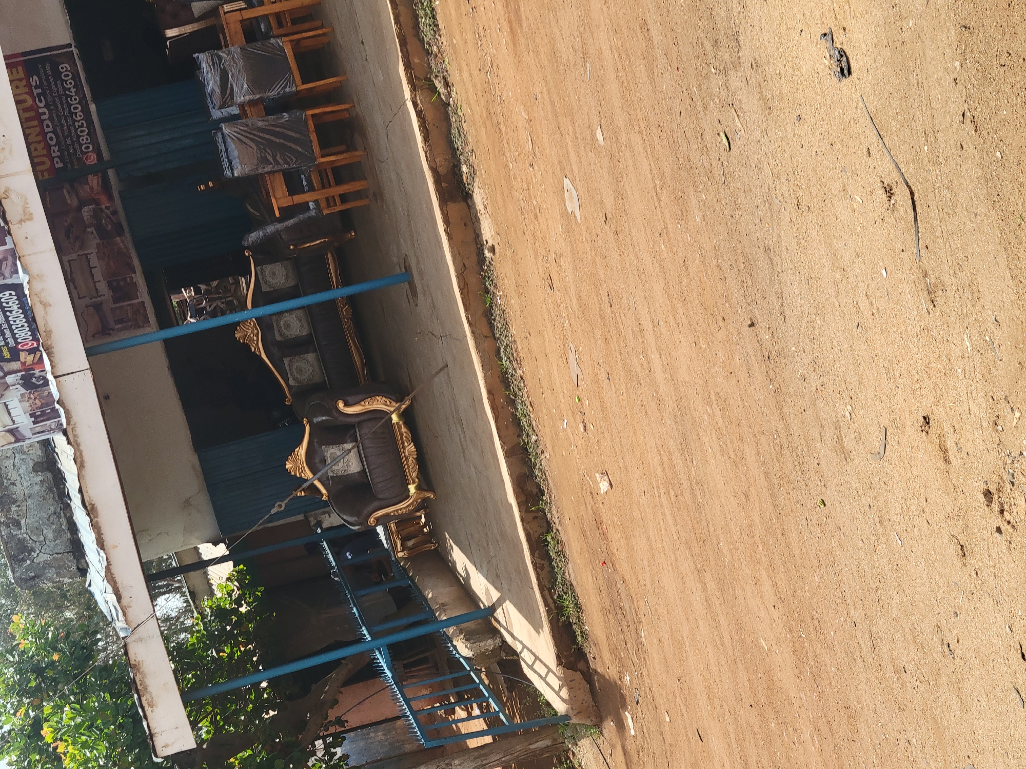 Here's a description of the image:
The image shows a furniture shop located on No 86, Yokubu Gowen Express Way, Dodin Kowo Jos Ploteau State. The shop displays various furniture items, including chairs with black covers and a sofa with ornate gold detailing. A sign for "FURNITURE PRODUCTS" with the phone number 08036064609 is visible. Outside the shop, there is a sandy area, likely the street or a sidewalk. The image is taken from an elevated angle and appears to be slightly rotated.