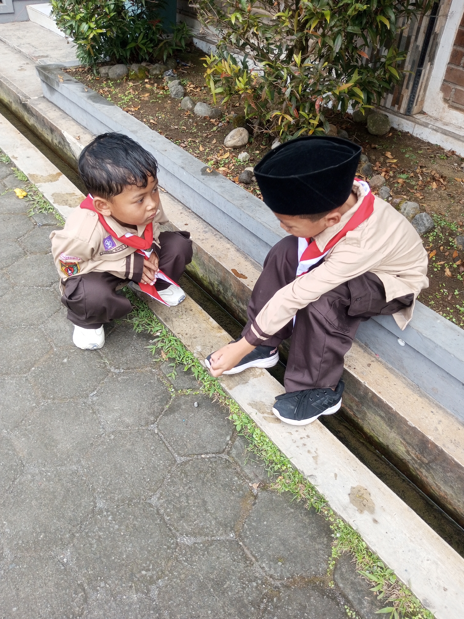 Two young boys, dressed in scout uniforms, are crouching on a concrete drainage channel. The boy on the left wears a light brown shirt with patches on the sleeve and a red and white scarf. He is looking down at his feet, wearing white shoes. The boy on the right wears a black peci hat, a light brown shirt with a red and white scarf, and dark brown pants. He is reaching down to adjust his black shoes. The background consists of lush green plants, rocks, and a brick wall, suggesting an outdoor setting. The ground is paved with interlocking hexagonal concrete tiles. This scene is located in Kalibeber, Indonesia.