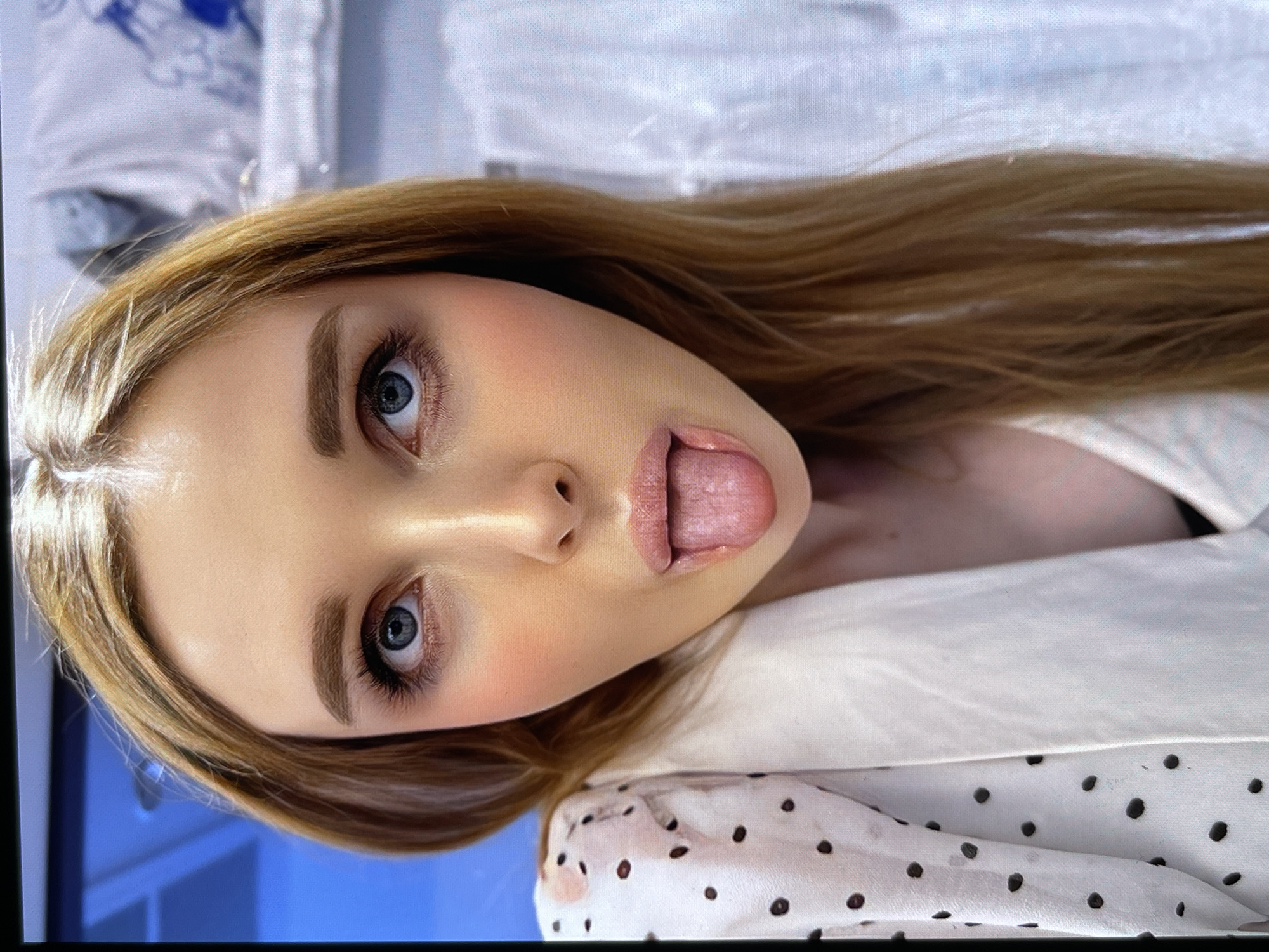A close-up, head-and-shoulders portrait shows a young Caucasian woman with long, reddish-blonde hair. She is looking directly at the camera with bright blue eyes and her tongue is slightly sticking out. Her makeup includes prominent eyelashes and rosy blush on her cheeks. She is wearing a white garment with dark polka dots and a white lapel or collar. The background is blurred and appears to be indoors, with hints of blue and white patterned fabric or surfaces. There are no visible environmental features to determine a specific city or location.