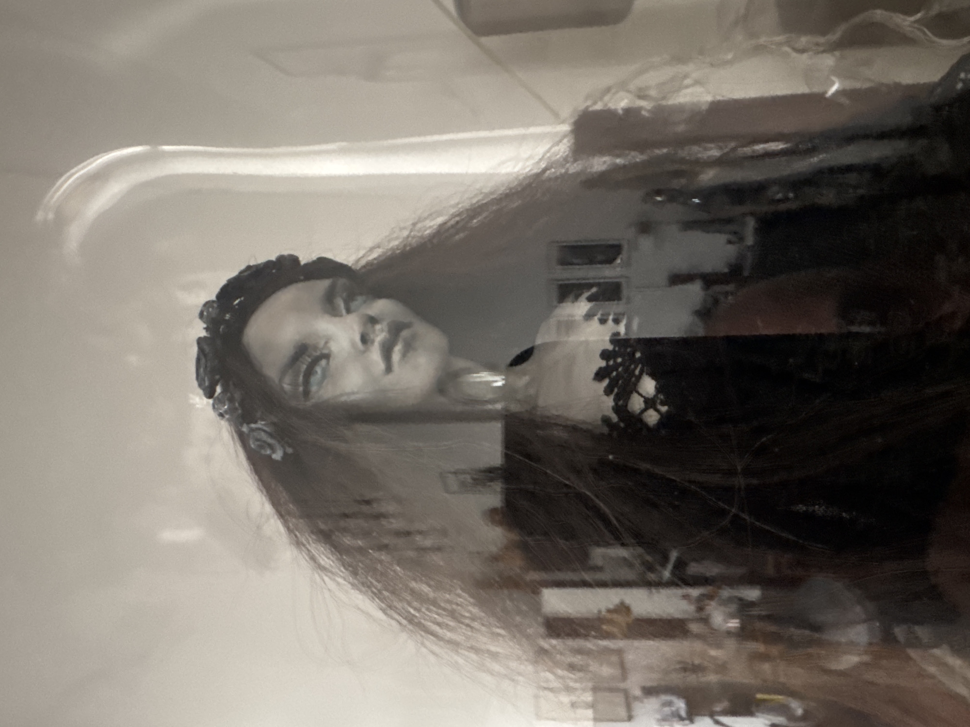 The image shows a close-up, slightly distorted, and reflected view of a doll or mannequin with long, dark hair and a floral headband. The doll has striking blue eyes and is wearing a black, lace-accented dress. The background is blurred and reflects what appears to be an indoor environment with furniture and possibly artwork on the walls. The overall lighting is soft and diffused. There is no discernible information within the image that would indicate a location in Frankfurt, Germany.