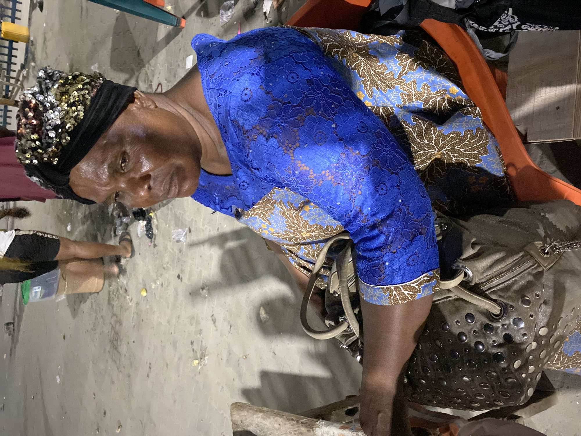 A medium-angle shot captures a middle-aged Black woman seated, likely in Lekki, Nigeria, given her attire and the general environment. She wears a blue lace blouse over a printed Ankara dress with brown leaf patterns. A sequined headwrap adorns her head. She sits on an orange chair with a gray studded handbag next to her. Debris litters the gray concrete floor, suggesting an outdoor or semi-outdoor location. Other individuals and objects are partially visible in the background, but are out of focus.