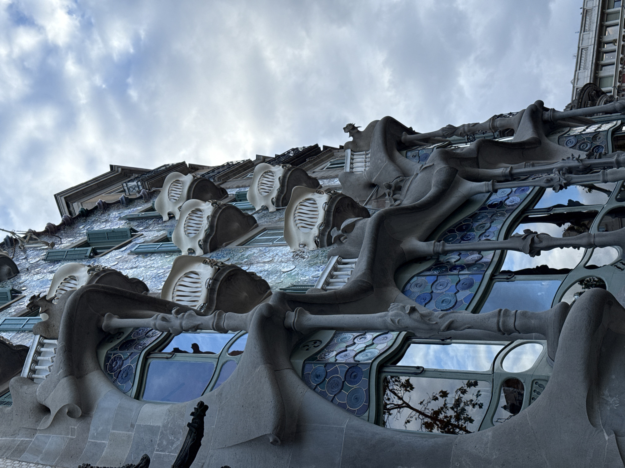 The image captures a low-angle, close-up view of Casa Batlló, a renowned building in Barcelona, Spain, designed by Antoni Gaudí. The building is characterized by its skeletal and organic architectural style. In the frame are various architectural elements, including wavy stone facades, bone-like balconies, and blue-toned mosaic window panes. The sky with light, cloudy patches is visible above the building. The image offers a ground-level perspective emphasizing the building's whimsical and unique structure.