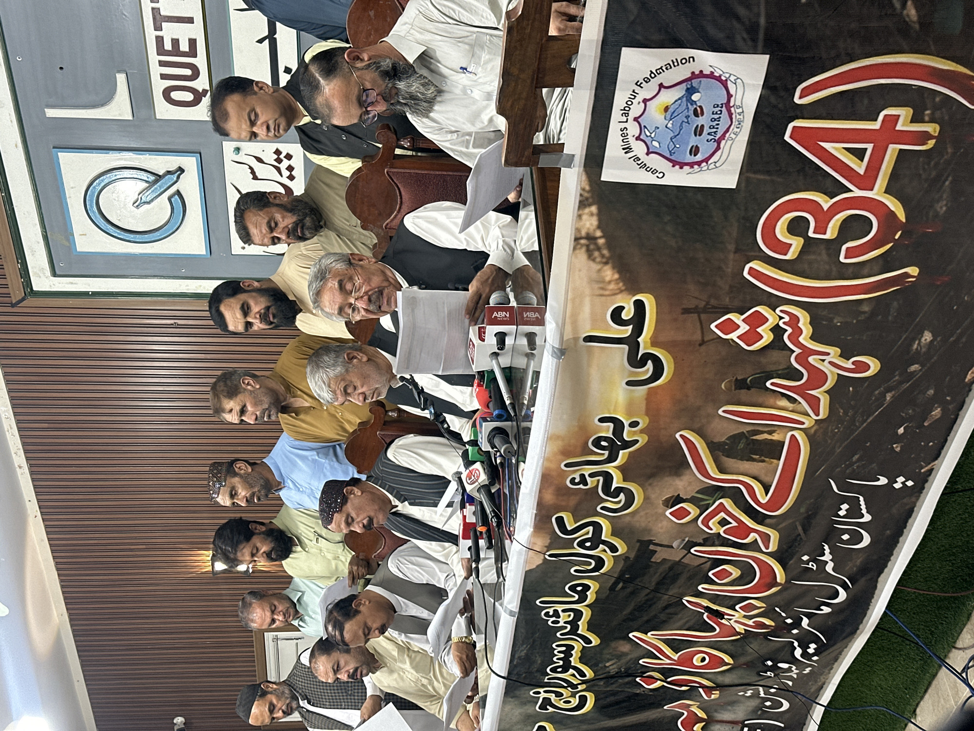 A group of approximately 15 men, most of whom appear to be middle-aged or older, are seated at a table in what seems to be a press conference or meeting. They are dressed in traditional Pakistani attire, including shalwar kameez and vests. Many of the men are holding papers, and microphones are positioned in front of them, suggesting they are speaking or about to speak. Behind them, a banner with Urdu text and imagery is visible. There is also a sign with a stylized "Q" and the word "QUETTA" in the background, indicating the location. The overall environment appears to be indoors, with wooden paneling on the walls and artificial lighting.