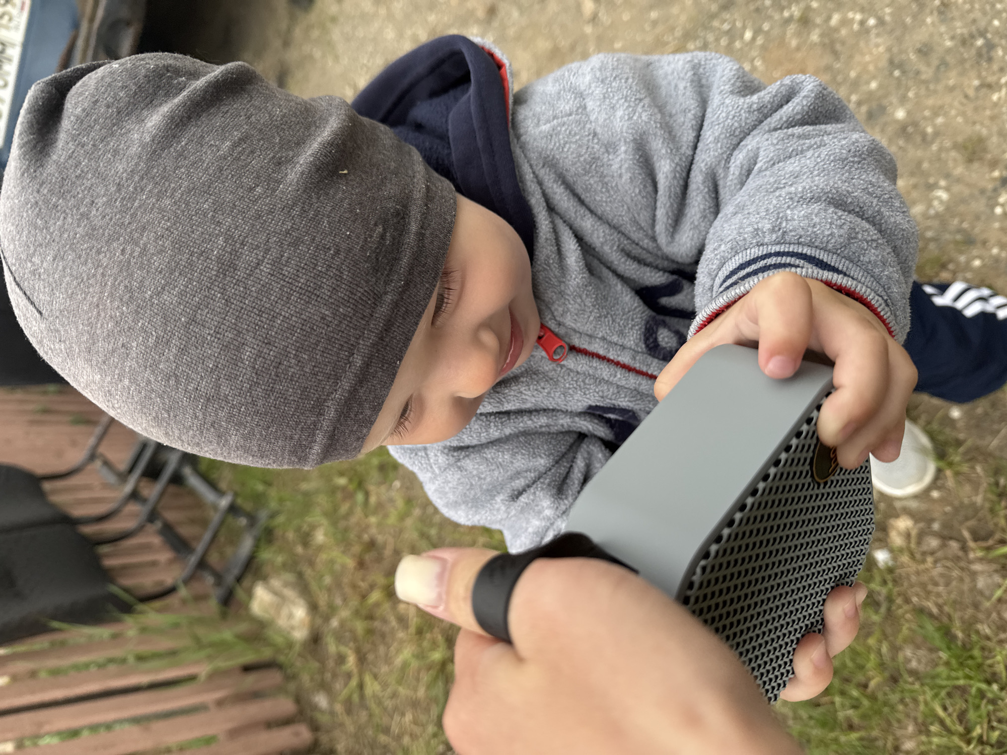 A high-angle outdoor shot captures a young child intently engaging with a small, grey rectangular electronic device, possibly a portable speaker. The child, likely a toddler, wears a grey knit beanie, a grey fleece jacket with a red zipper pull, and a dark blue hooded top underneath. They are looking down at the device with a gentle smile, holding it with both hands. The device features a perforated speaker grille on its visible side. In the immediate foreground, an adult's hand, with a long thumbnail and a black ring on the thumb, is also seen, either presenting the object to the child or holding it jointly.
The scene is set outdoors during daylight, under soft or overcast lighting conditions. The ground beneath is a mix of dirt and dry grass. In the blurred background, elements like brown wooden slats (possibly a fence or bench) and a dark metal structure are visible. The child's attire also includes dark pants with white stripes on the leg. A blurry white rectangle with unreadable dark text, resembling a license plate, is faintly discernible in the upper left background. The overall impression is one of gentle interaction and discovery, situated in an outdoor environment in Vereshchagino, Russia.