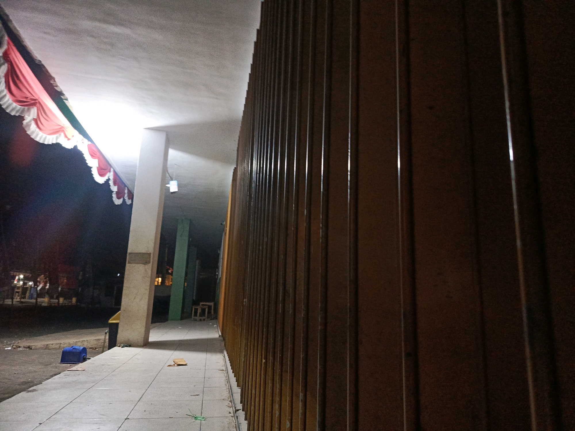 An outdoor, low-light, wide-angle photograph captures a walkway and building facade in Pamekasan, Indonesia. The perspective is from ground level, looking down a tiled walkway that runs alongside a building with a corrugated metal wall. The wall dominates the right side of the frame and is a dark brown color, with vertical corrugated lines that create shadows and highlights.
To the left of the walkway, there is a light-colored concrete pillar. Above and behind the pillar, a bright light source illuminates a section of the ceiling and casts a strong light onto the walkway. A decorative red and white valance, possibly part of a banner or curtain, is draped along the top edge of the illuminated area.
Further down the walkway, a blue plastic bin is visible on the ground, near the edge of the walkway. Beyond the walkway, there is an unpaved area with some debris and what appear to be vehicles or stalls in the distance, suggesting a public or commercial space. On the walkway itself, there are a few scattered pieces of debris, including a light brown object.
In the background, between the concrete pillar and the corrugated wall, the walkway continues, leading to a green-colored pillar and some wooden stools. The overall scene suggests a nighttime or dusk setting, with artificial lighting being the primary source of illumination.