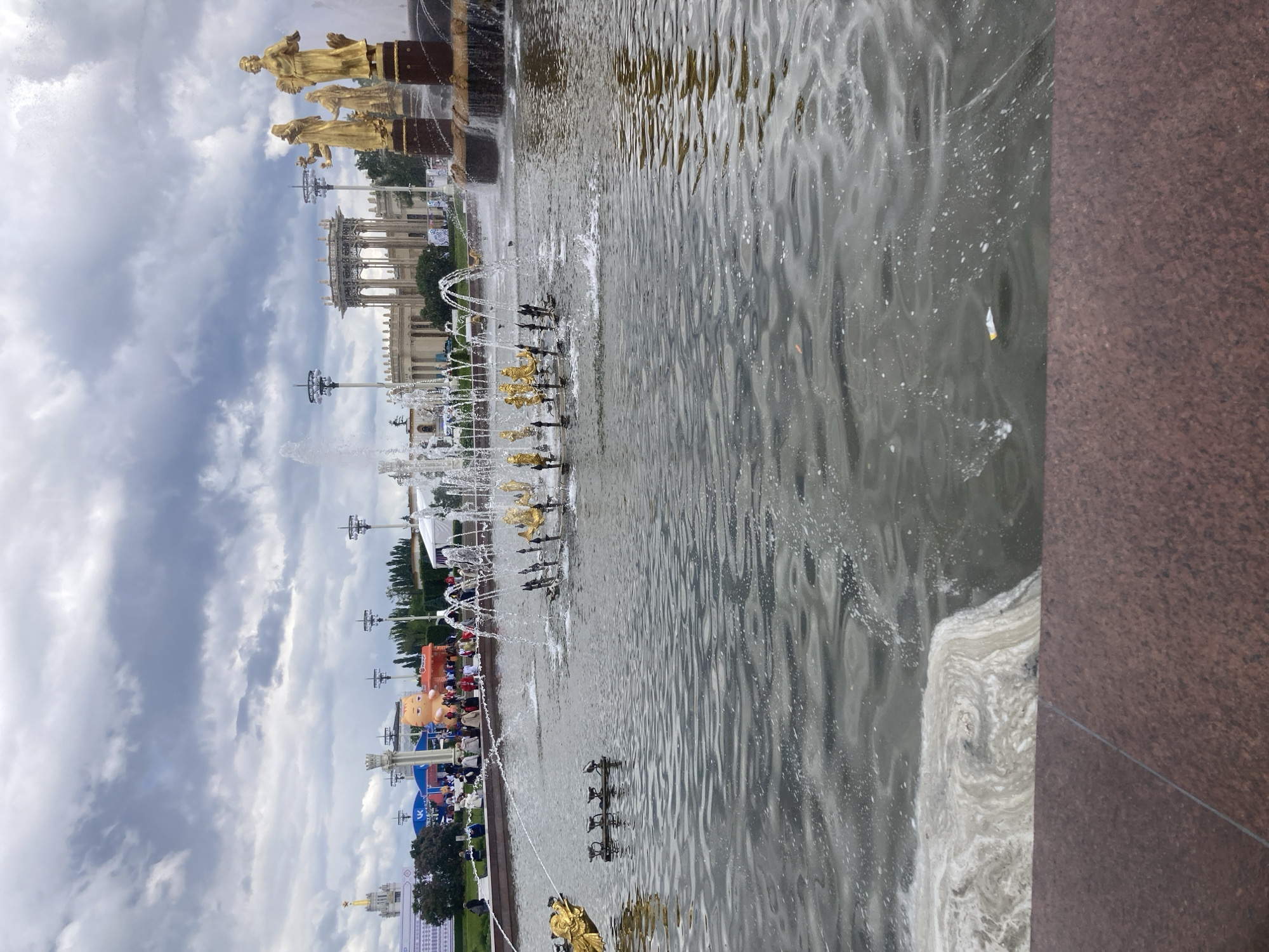 The image captures a view of the Friendship of Nations Fountain at the All-Russia Exhibition Center (VDNKh) in Moscow, Russia. The fountain features golden statues and water jets. In the background, various pavilions and structures of VDNKh can be seen under a cloudy sky. People appear to be gathered in the distance. A part of the fountain basin and its edge are visible in the foreground.