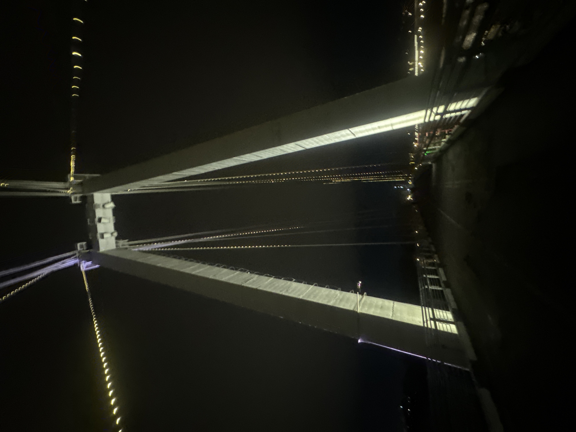 The image shows a close-up, nighttime view of the Yenisei River Bridge in Krasnoyarsk, Russia. The bridge is a suspension bridge, and the image shows the towers, the cables, and the bridge deck. The towers are illuminated, as are the cables, creating a bright, geometric pattern against the dark sky. The deck appears illuminated with smaller lights. The environment is dark, suggesting it is nighttime. There is no indication of people, subjects, objects, actions, or interactions, other than what is inherent in the scene.