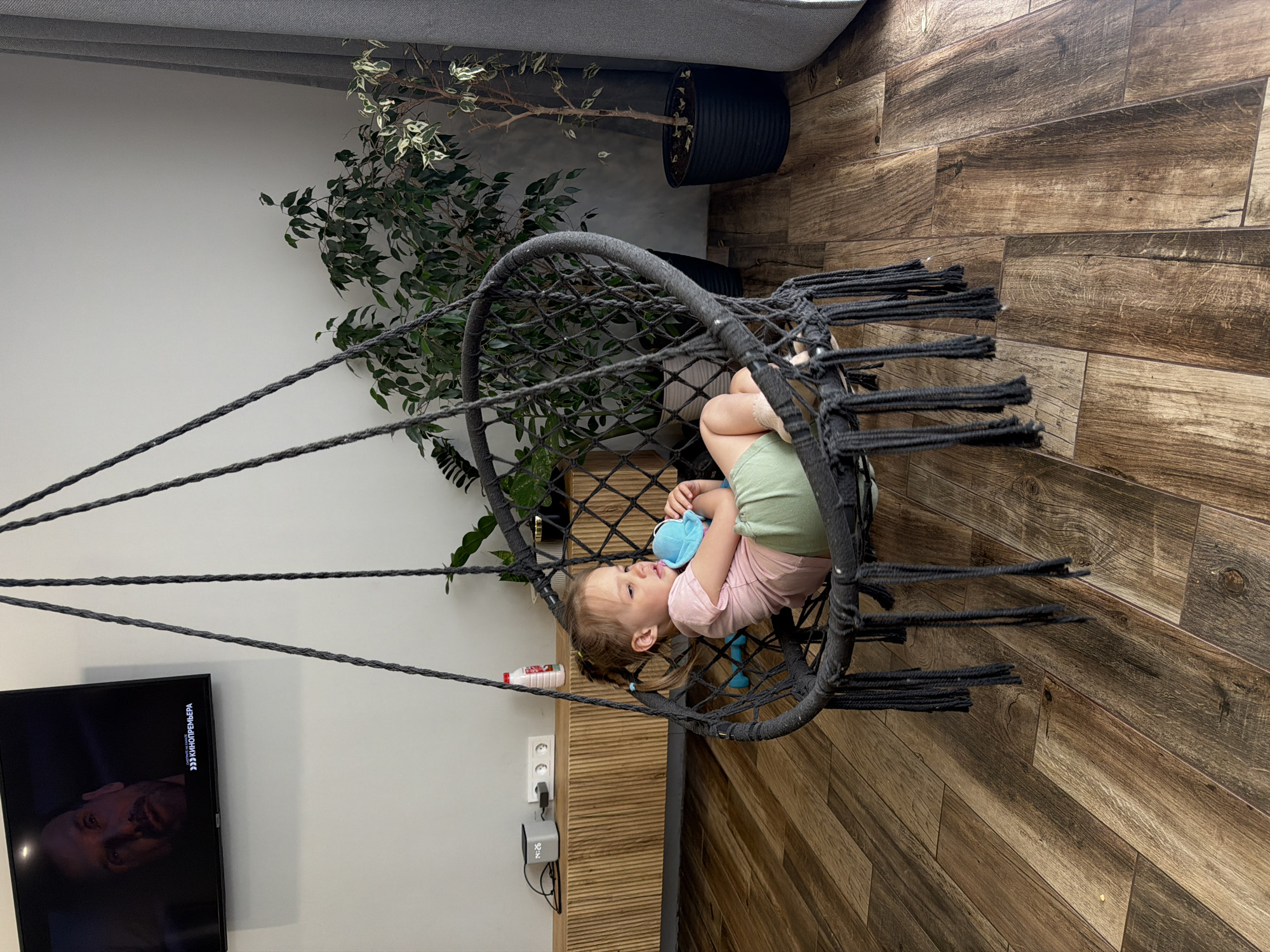 A child reclines in a black macrame hanging chair, cradling a blue stuffed animal. The chair is positioned indoors on a brown wood floor, next to a cabinet with a white outlet and a gray temperature control device. A red and white bottle sits on the cabinet's surface. To the left, a flat-screen TV displays a man's face on screen with Russian text in the top right corner. Behind the chair are potted plants with green foliage. Based on the text on the TV and information provided, the location is Chaikovsky, Russia.