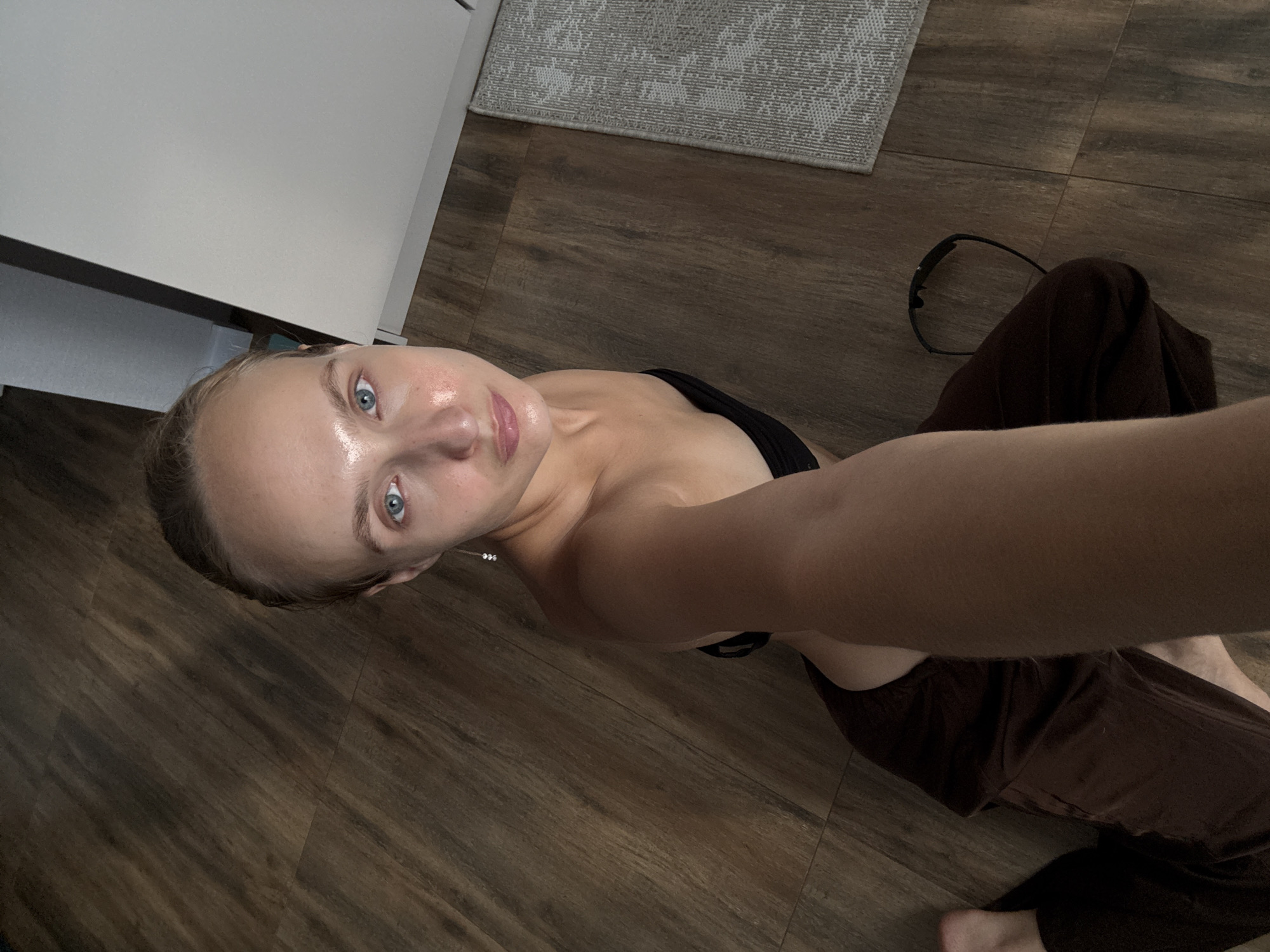The image shows a young woman lying on a wooden floor. She is looking up at the camera with a slight smile. She is wearing a black top and brown pants. Her skin is dewy, suggesting she may have just applied skincare or is feeling warm. The setting appears to be indoors, with a white cabinet and a grey rug visible in the background. There are no visible text or any specific indications of time of day or weather. The overall mood is relaxed and casual.