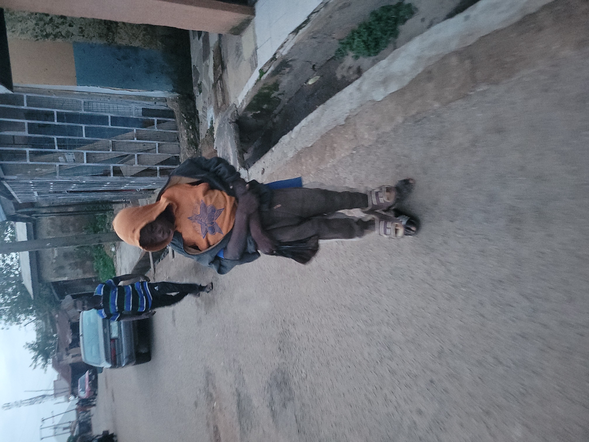 In Jos, Nigeria, a street scene unfolds on an overcast day. A young person, wearing an orange hooded sweatshirt over a dark shirt, dark pants, and sandals, walks towards the viewer. They are carrying a black bag over their right shoulder. To the left and further down the street, a man in a striped blue, black, and white shirt and dark pants walks away from the viewer, his back to the camera. A dark-colored sedan is parked on the left side of the road. Buildings line the right side of the street, one of which features a metallic security grate over its windows. A concrete drainage ditch runs alongside the street on the right. The overall atmosphere is muted, suggesting the time of day may be late afternoon or early morning.