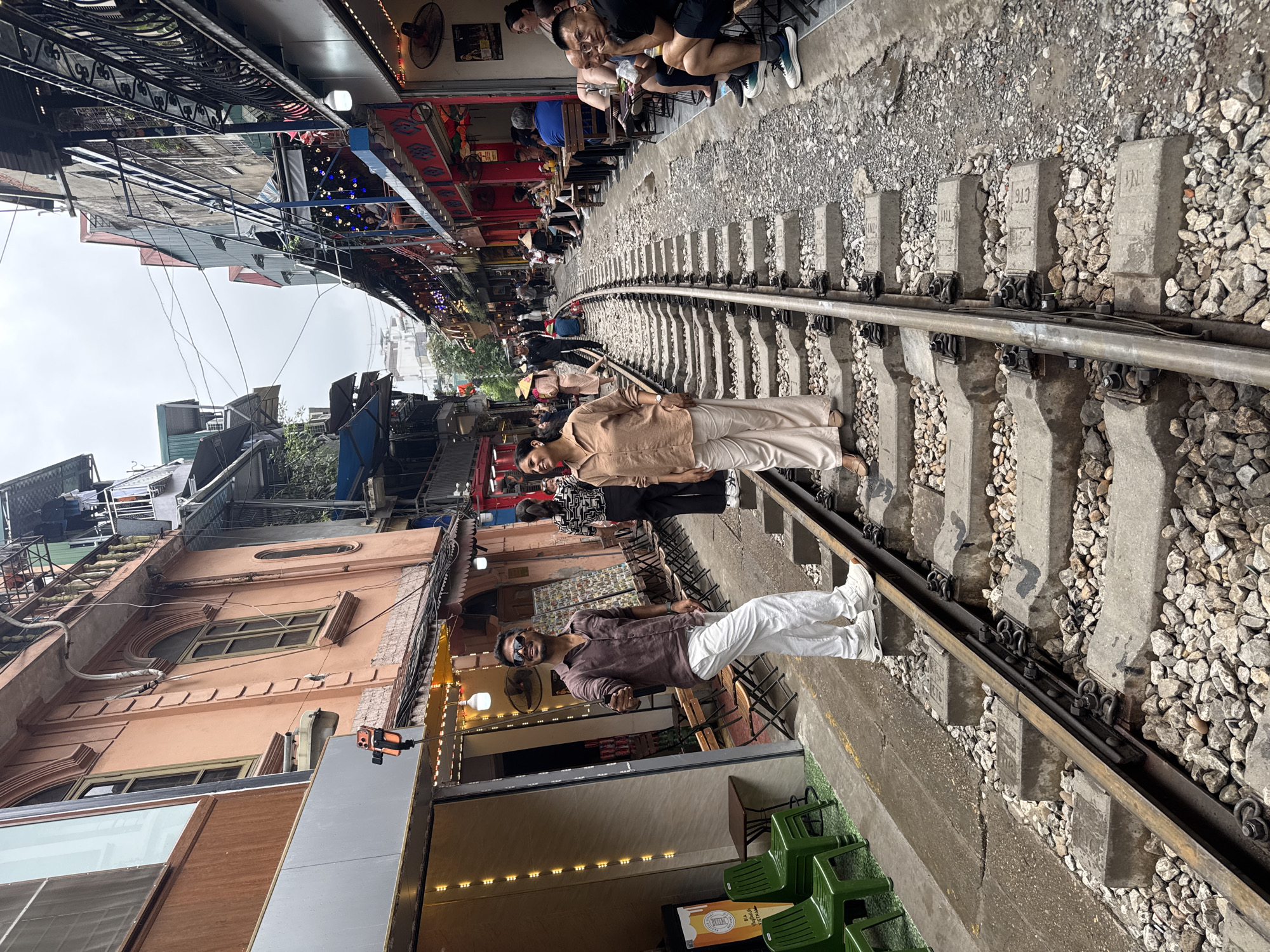 The image shows a section of the Hanoi Train Street in Vietnam. A railway track runs through a narrow alley with buildings closely bordering it on both sides. People are standing on the tracks, posing for photos. Some are seated at outdoor cafes alongside the railway. The setting appears to be a tourist attraction.