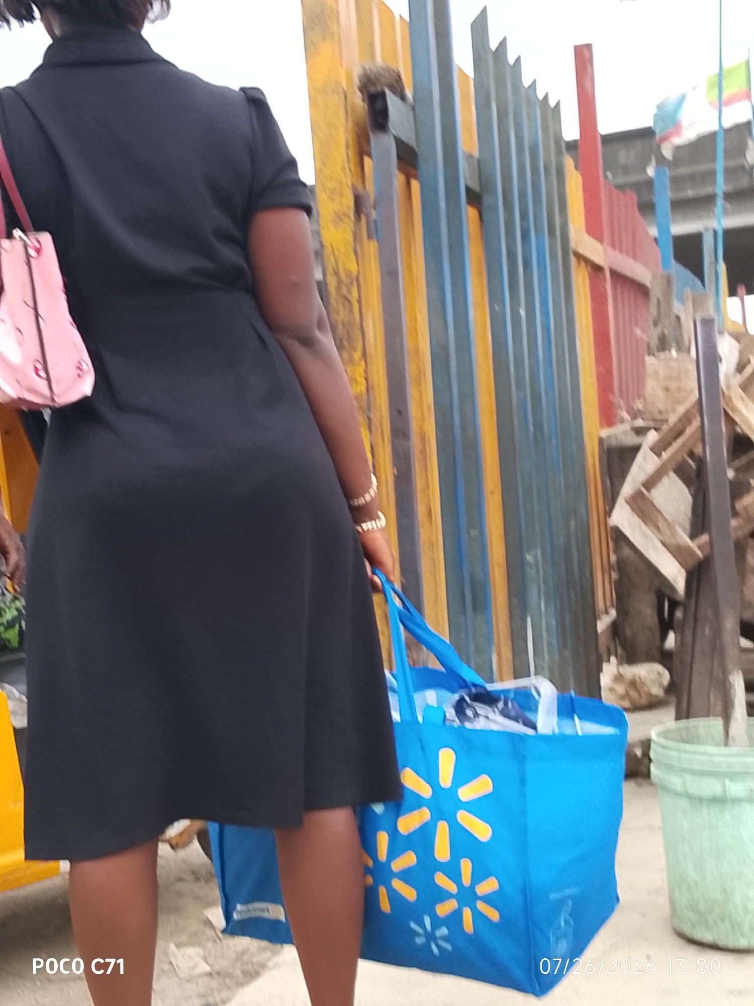 In Lekki, Nigeria, a woman wearing a black dress stands with her back to the camera, holding a pink handbag on her left shoulder and a blue reusable shopping bag in her right hand. The shopping bag is adorned with yellow starburst patterns, characteristic of Walmart branding. She is positioned in front of a colorful corrugated metal fence, composed of vertical panels painted in yellow, blue, and red. To the right of the woman, a green plastic bucket is visible. The ground beneath them appears to be a mix of sand and debris, with some discarded wooden planks and debris in the background. A timestamp in the lower right corner indicates the date and time as 07/26/2026 13:00, and a watermark in the lower left corner reads "POCO C71".