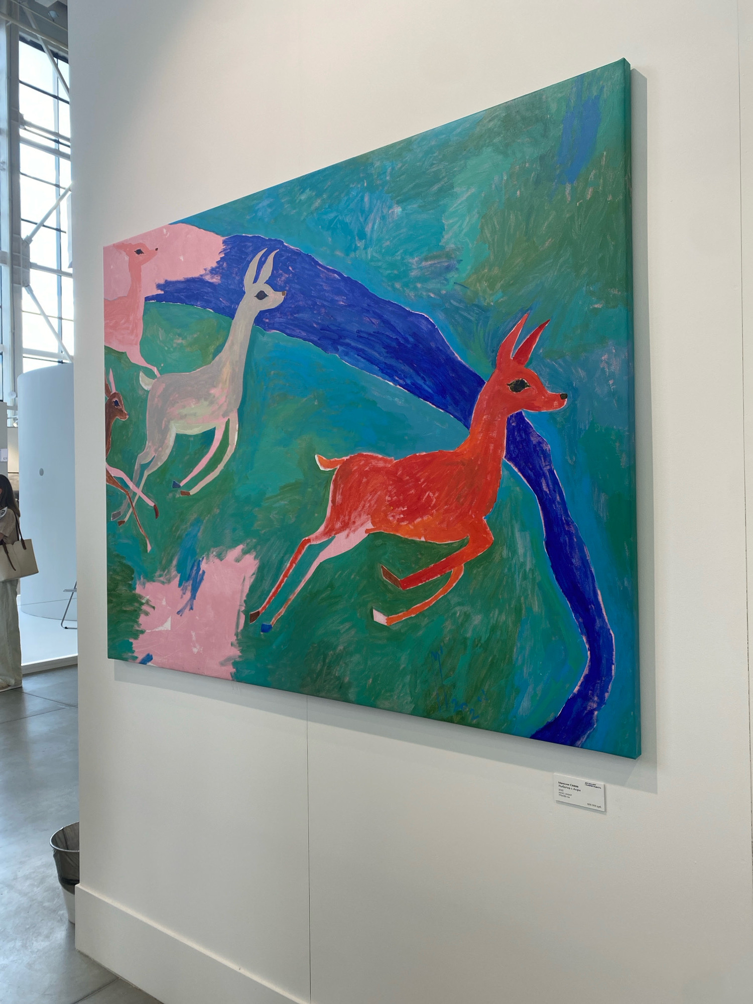 The image depicts a large, horizontal painting of several deer rendered in a somewhat abstract and expressive style. The dominant colors are various shades of green, blue, and pink, with a prominent red deer in the foreground. The background appears to be a stylized landscape with a blue, winding element that could represent a river or a path. The painting is hung on a white wall in what appears to be an art exhibition or gallery space. To the left of the painting, there's a glimpse of a woman in light-colored clothing, carrying a large tote bag. The environment seems to be an indoor public space with polished concrete floors and large windows with metal framing in the background. A metal trash can is visible on the floor near the bottom left of the frame. A small label is attached to the wall beneath the painting, indicating it is an artwork for sale. The overall impression is that of an art exhibition in a contemporary, well-lit space. The image is framed from a slight angle, showcasing the painting and its immediate surroundings.