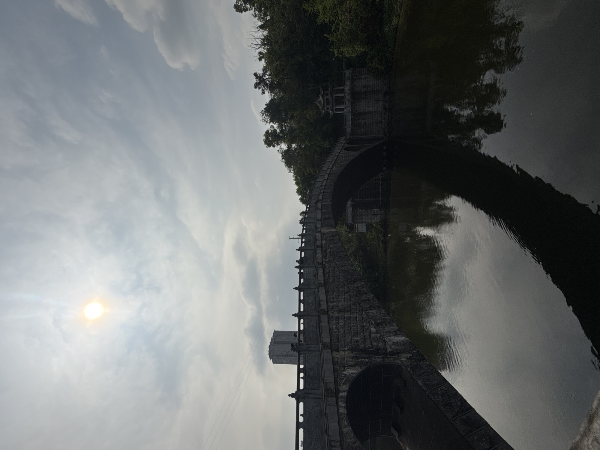 The image shows a stone bridge spanning a dark body of water, likely a river or canal. The bridge is constructed with arched openings and a decorative railing. On the far side of the bridge, there's vegetation and a small building. A modern building can be seen rising near the middle of the bridge. The sky is cloudy with the sun shining through. The image appears to be taken in Jiulongpo District, China, but this is only based on information provided and cannot be confirmed.