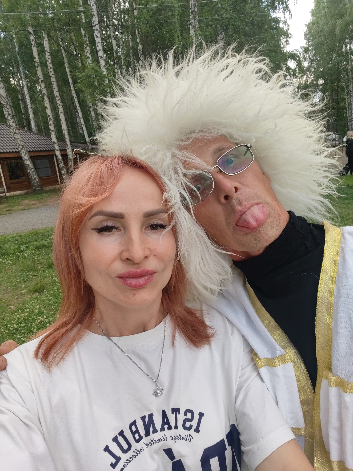 This is a close-up, selfie-style medium shot of a man and a woman posing outdoors in a wooded area, likely in Chaikovsky, Russia. The man, on the right, is wearing a large, fluffy white hat and glasses, and he is sticking out his tongue playfully. He is also wearing a black turtleneck and a white garment with gold trim. The woman, on the left, has shoulder-length, reddish-blonde hair and is wearing a white t-shirt and a silver necklace with a crown pendant. The background consists of birch trees, a small wooden building, and green grass with white flowers. The lighting suggests it is daytime, possibly late afternoon or early evening, with overcast or diffused light. The overall mood appears lighthearted and fun. The woman's t-shirt has dark blue text that reads "ISTANBUL" in large block letters, with smaller text underneath.