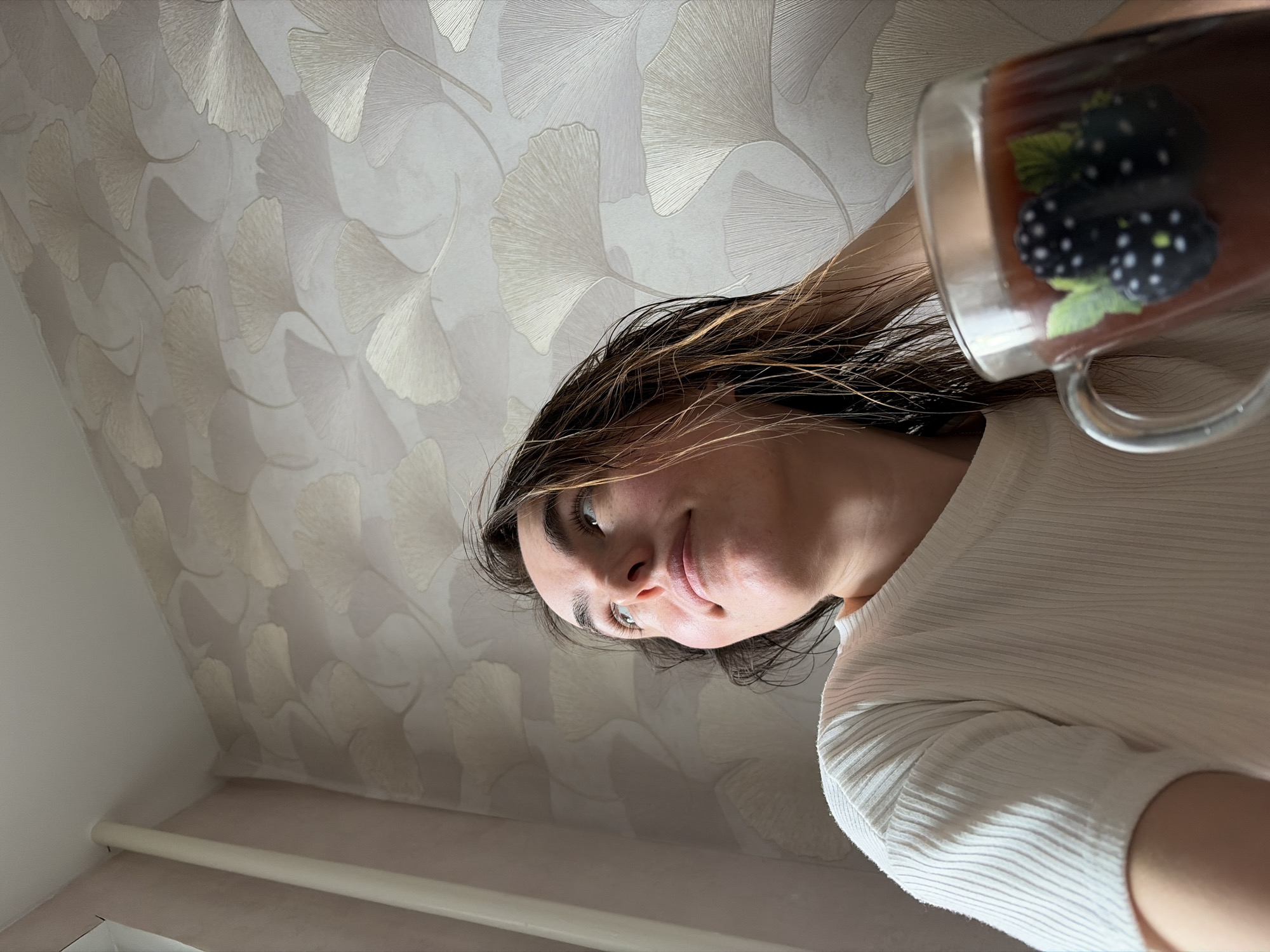 The media file captures a young woman from a low-angle perspective, giving the impression of looking up at her. She has long, dark hair with visible lighter streaks, appearing slightly damp or textured. Her light-colored eyes are open, and she wears a gentle, pleasant expression with a subtle smile. She is dressed in a light-colored, possibly off-white or cream, ribbed long-sleeved top.
In her right hand, partially visible on the right side of the frame, she holds a clear glass mug containing a dark liquid, likely tea or coffee. The mug is decorated with an illustration of two blackberries and green leaves.
The background features a wall covered in light-toned wallpaper, patterned with stylized ginkgo leaves in beige or gold hues against a lighter, possibly off-white, backdrop. Below the patterned wallpaper, a light-colored skirting board or trim runs horizontally along the lower part of the wall, with a plain light-colored wall visible in the bottom left, suggesting an interior corner. The image's composition appears to be rotated counter-clockwise from a standard upright view. The scene depicts a casual indoor moment, likely at home, in Izhevsk, Russia.