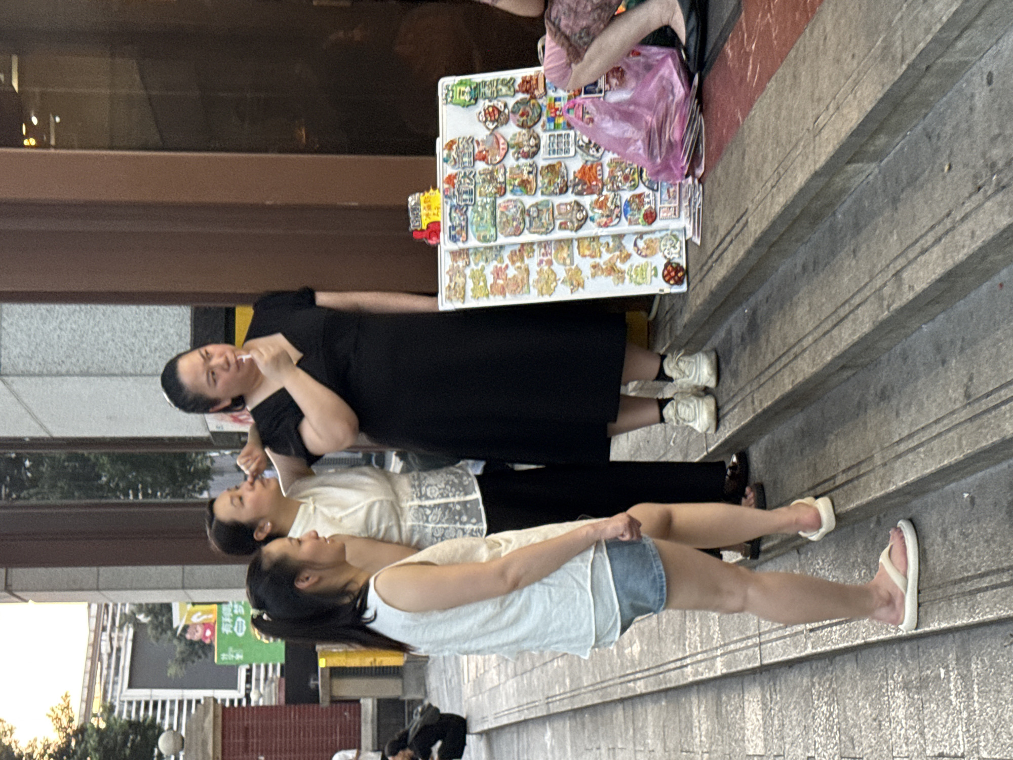 Three women are standing on a set of outdoor stairs in Yuzhong District, China. The woman on the left is wearing a white sleeveless top and denim shorts. The woman in the middle is wearing a white collared shirt with decorative patterns and black pants. The woman on the right is wearing a black dress and holding a white object to her mouth, possibly a cigarette or candy. In front of the woman on the right are two white boards displaying various colorful stickers or magnets, resembling souvenirs. A pink plastic bag is placed on the stairs next to the display boards. The background shows a building facade with a dark brown entrance and a glimpse of trees and other structures further away. The lighting suggests it is daytime, possibly late afternoon, with sunlight casting shadows.