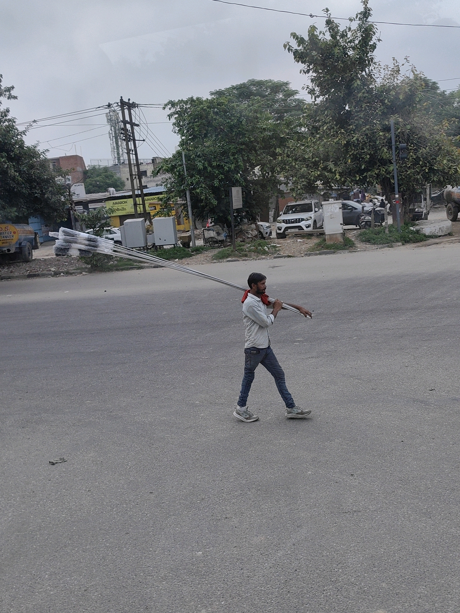 A man is walking on a road in Raipur Khurd, India, carrying metal poles over his shoulder. The man is wearing a light blue patterned shirt, dark blue jeans, and grey athletic shoes. He has a red cloth draped around his neck. He is walking from left to right in the frame, and the poles he is carrying are wrapped in plastic. In the background, there are trees, buildings, parked cars, and a power pole with wires. The sky is overcast.