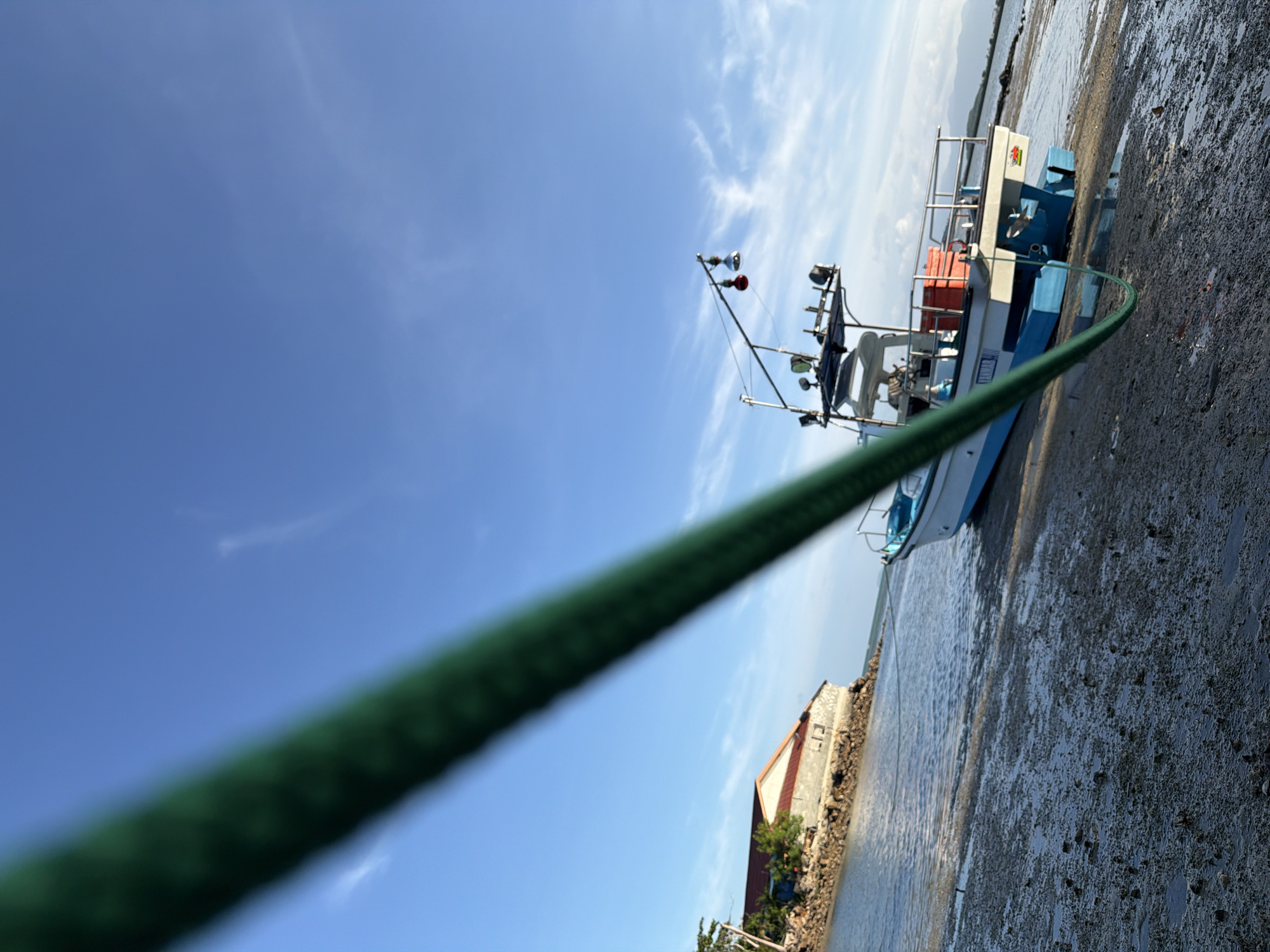 A low-angle, Dutch-tilted shot captures a white and blue boat resting on a muddy, rocky shore under a clear blue sky with scattered white clouds. A thick, green rope diagonally bisects the foreground, obscuring part of the sky and the boat. The boat, positioned on the right side of the frame, has a cabin with windows and a metal railing around its deck. Two red boxes are visible on the deck. In the background, on the left, a low, rectangular building with a reddish-brown roof sits on a rocky embankment. The water is calm, and the environment suggests a coastal or harbor area.