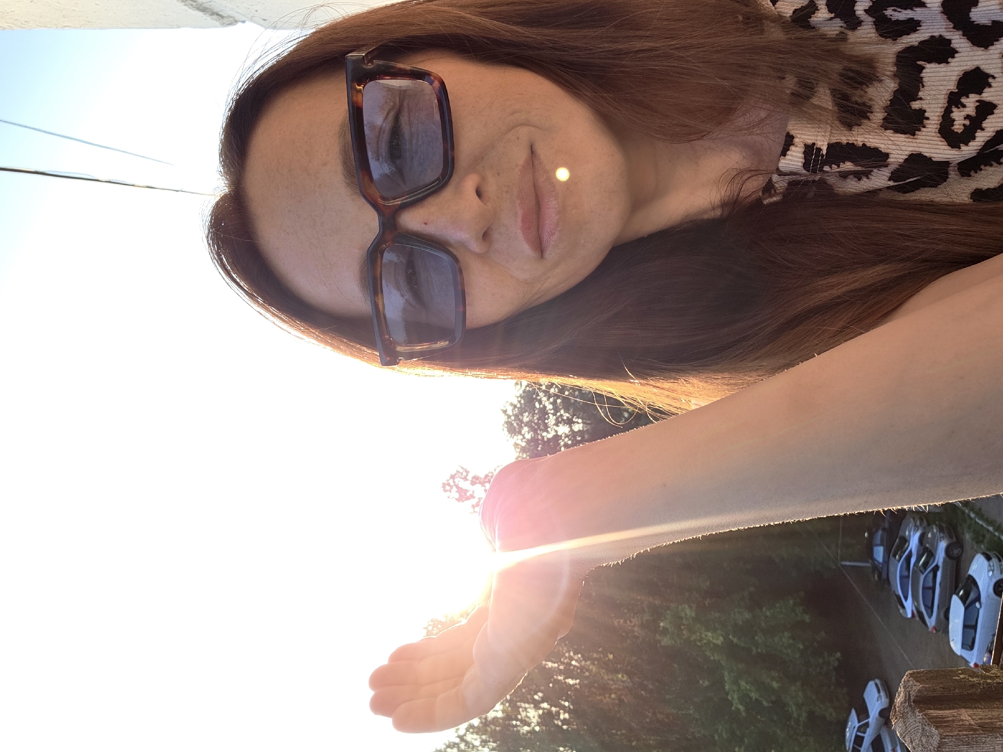 A woman with long brown hair and sunglasses is holding her hand up to block the sun. The sun is shining brightly, creating lens flare and a halo effect on her hand. She is wearing a leopard-print top. In the background, there are trees and a parking lot with several cars. The scene appears to be outdoors, possibly on a balcony or rooftop, with a clear sky. The image was taken in the city of Chaikovsky, Russia.