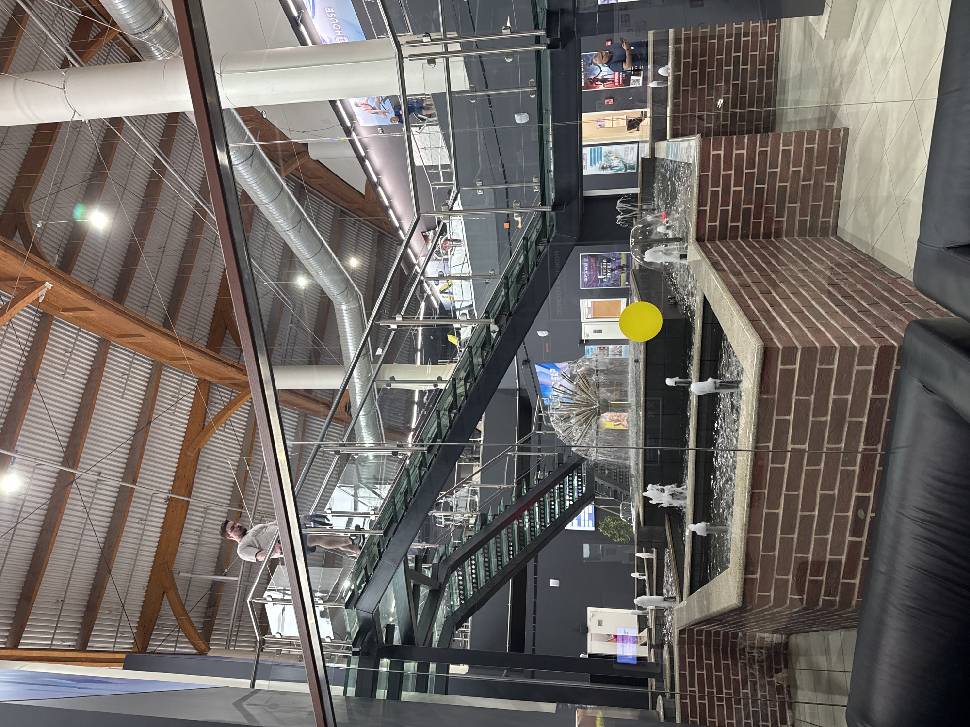 The image depicts an interior scene, seemingly a modern building or a shopping center. A multi-tiered water fountain with brick borders is situated in the foreground. Water spouts rise from each level of the fountain. Behind the fountain, a black staircase with glass railings leads to upper levels. There are advertisements or posters on the wall behind the staircase. People can be seen on an upper level. The ceiling has exposed wooden beams and metal ductwork. A yellow circle is partially obscuring the view of a sign. The exact location in Saint Petersburg, Russia is not clearly identifiable from the image. The scene does not show any clear actions or interactions beyond the water fountain's operation and people occupying the space. The environment appears modern and well-lit.