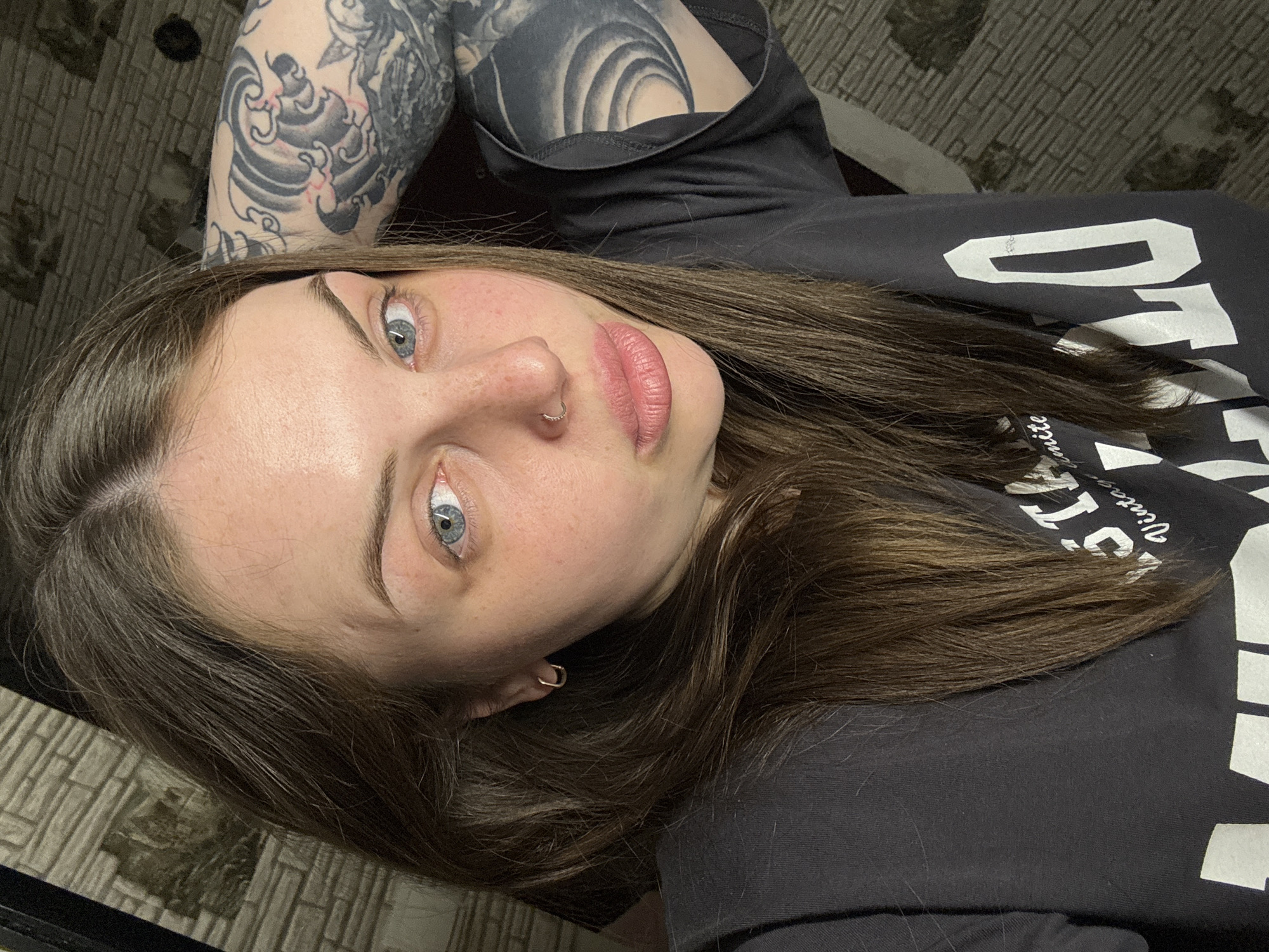 A young woman with light skin, blue eyes, and brown hair is lying down. She has a nose ring and small hoop earrings. A portion of her upper left arm is visible, showing a black and grey tattoo. She is wearing a black t-shirt with white lettering. The background shows a tiled surface and a patterned blanket. The location is Chornomorskoe, Ukraine. No specific actions or interactions are observable.