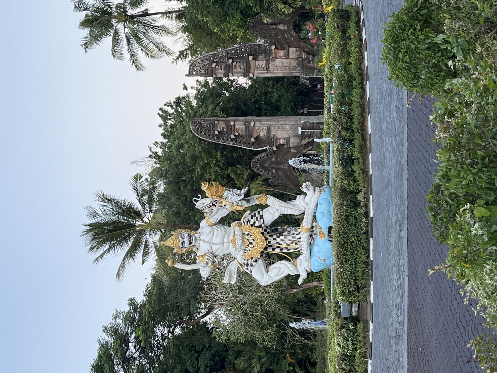 The image depicts a statue of a male figure riding a dragon-like creature, located in Unknown, Indonesia. The statue is white and gold, with checkered patterns on the figure's clothing. A palm tree is visible behind the statue. In the background, there is a split gateway structure made of stone. A fountain is located near the statue. Lush green vegetation surrounds the statue and gateway. A grey brick pathway is seen alongside manicured bushes. The sky is light blue and clear.