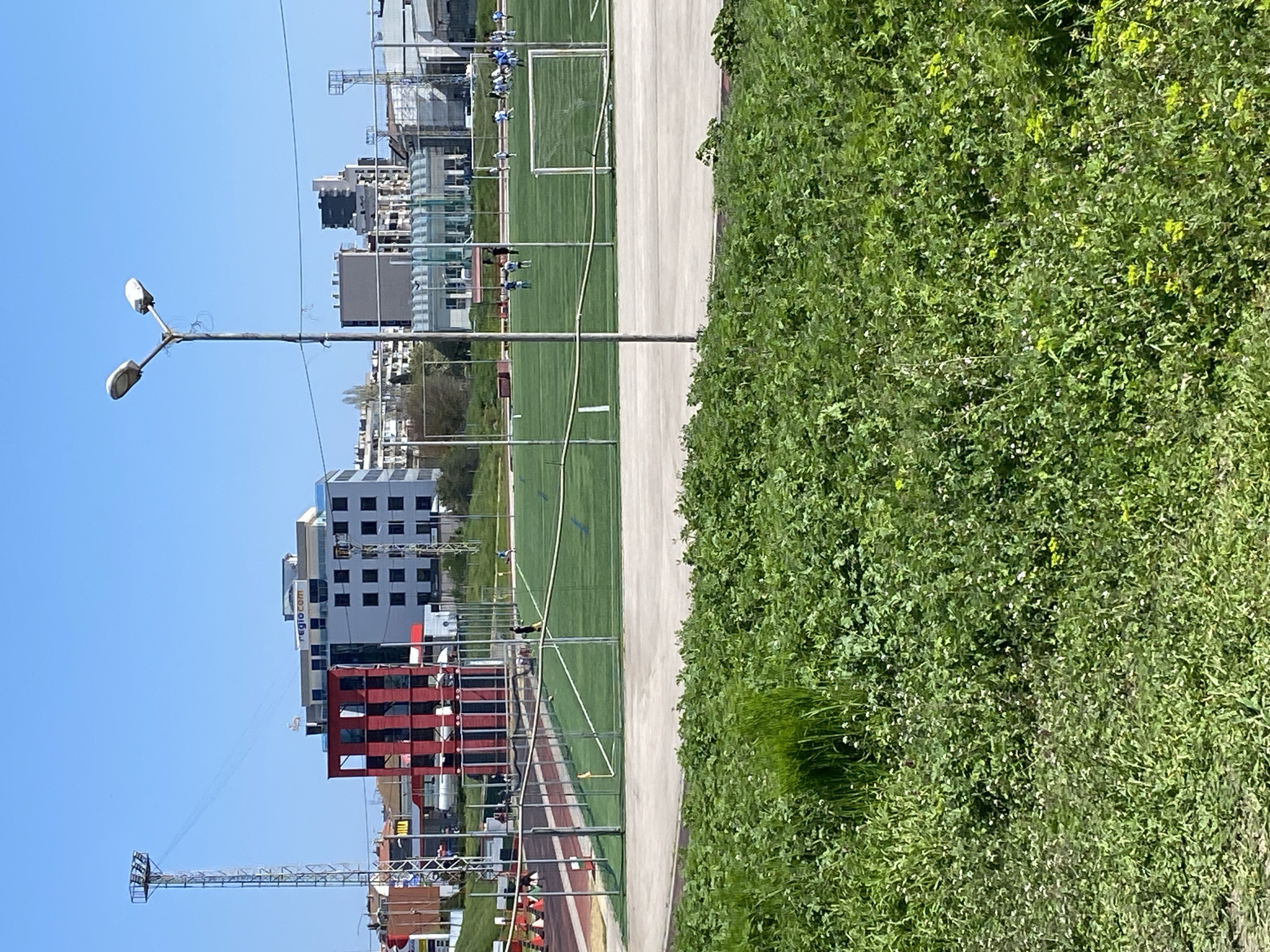 The image depicts a sports complex in Varna, Bulgaria, featuring a track and field area with red running lanes and orange cones. Adjacent to the track is a green artificial turf field with soccer goals. A concrete pathway separates the field from a dense patch of greenery. Beyond the sports complex, there are several buildings of varying heights and architectural styles, including one with red accents and another with a "regio.com" sign. Overhead power lines stretch across the scene, and a tall metal tower stands to the left. A street lamp is also visible. The sky is clear and blue. People can be seen on the track area.