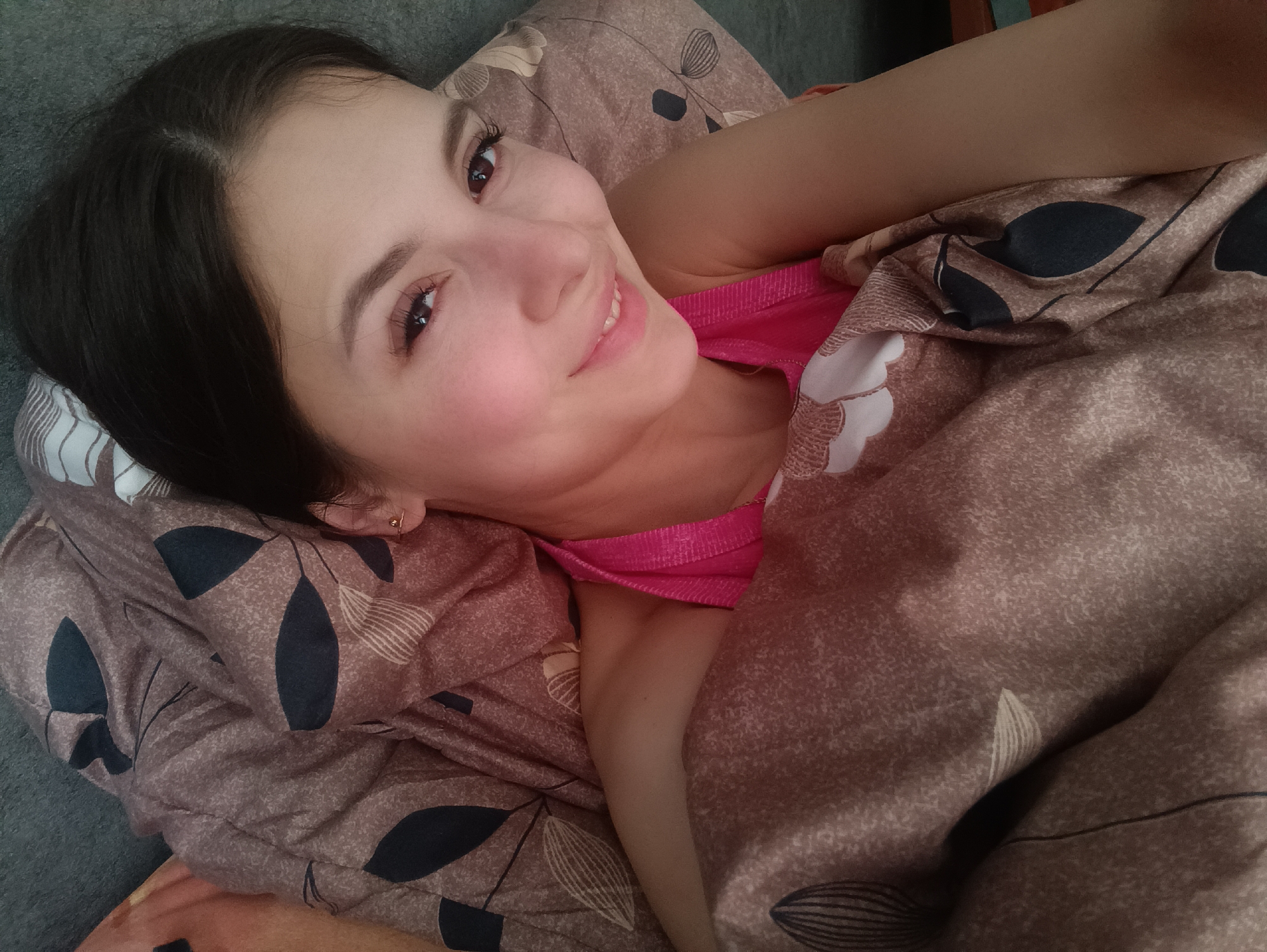 A young woman is depicted lying down, smiling brightly at the camera, in what appears to be a bed. She has dark hair, which is parted on the left side, and dark eyes with long eyelashes. Her expression is cheerful and relaxed, with a wide smile that shows her teeth, and a rosy blush on her cheeks. She is wearing a vibrant pink sleeveless top or camisole.
She is partially covered by a duvet or blanket, which features a repeating pattern of dark, stylized leaves on a light brown or taupe background. A pillow with a similar pattern is visible behind her head. The background behind her and the bed is a dark gray color, possibly a headboard or wall. The image appears to be a selfie taken from an overhead angle, looking down at the woman. The setting is clearly indoors, likely a bedroom, given the presence of bedding. No specific location cues, time of day, or visible text are discernible.