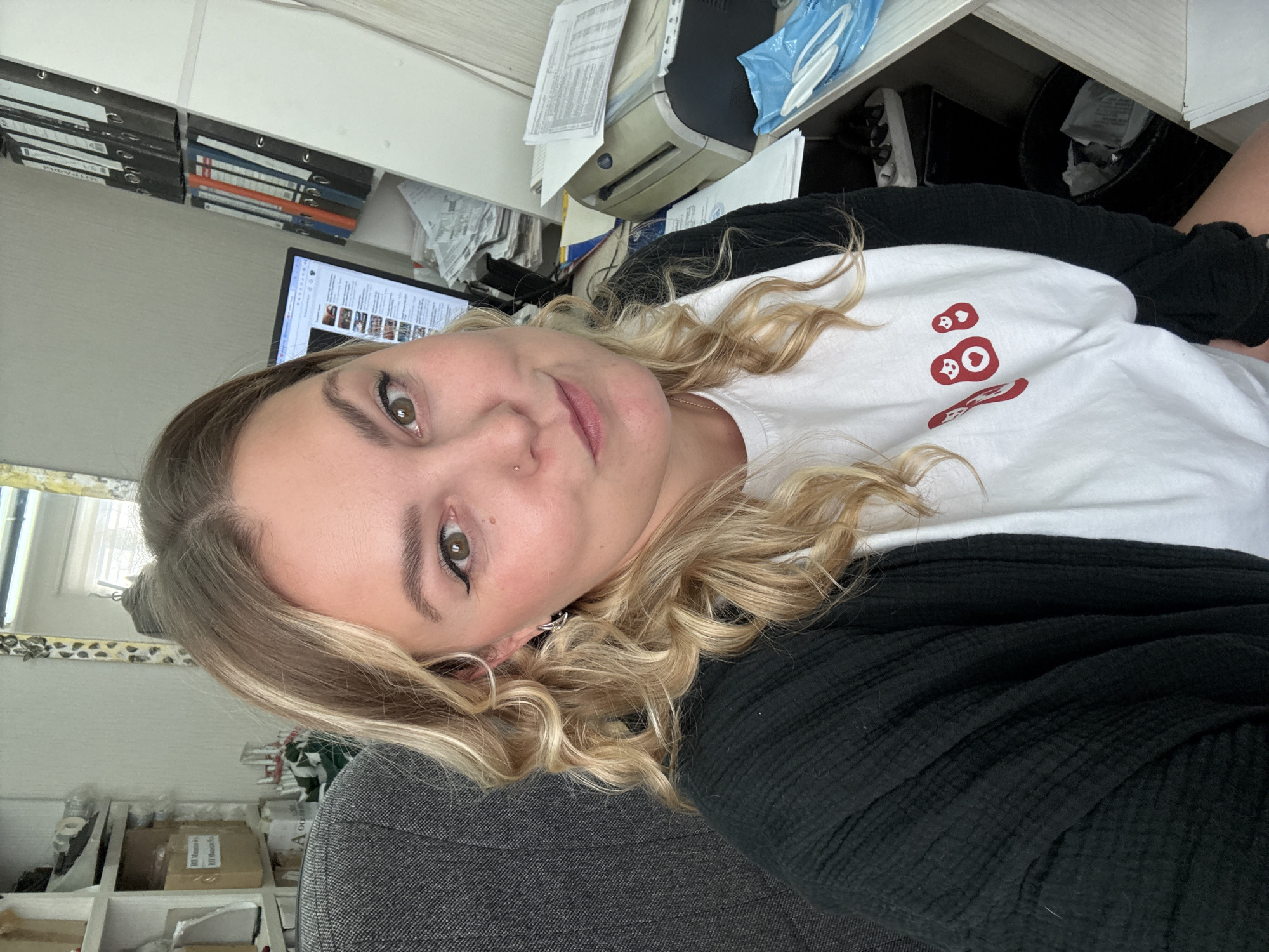 A Caucasian woman with blonde, curly hair is shown from the chest up, seated in an office. She is looking at the camera with a slight smile and her head tilted. She is wearing a white t-shirt with red nesting dolls printed on it, and a black cardigan over the t-shirt. She has makeup on, including eyeliner and mascara. Her hair is styled in loose curls.
The background of the image suggests an office environment. To the left, a grey office chair is visible. Behind the woman and to her right, shelves are filled with binders and boxes. A computer monitor is also visible, displaying what appears to be a webpage with thumbnail images. Further to the right, there is a printer and stacks of papers. A blue plastic bag containing white items is on a shelf in the foreground. A trash can is visible in the lower right corner. The lighting appears to be from an overhead source, creating some shadows.
The location is identified as the city of Noviy, Russia, based on the prompt. However, there are no specific visual cues in the image that definitively confirm this location. The scene depicts a typical office setting that could be found in many urban environments.