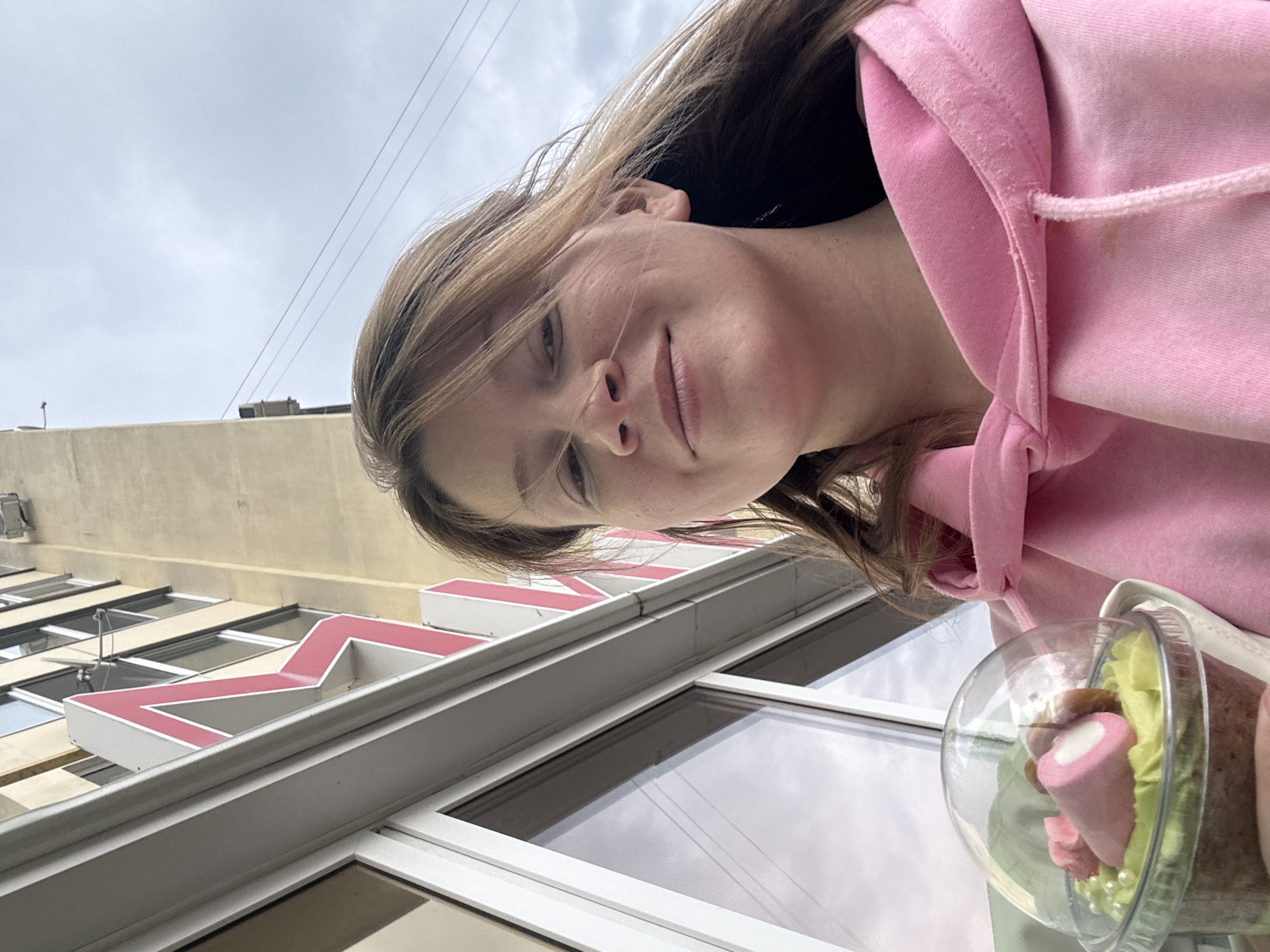 A young woman is featured in a close-up, slightly tilted photo taken outdoors in Chaikovsky, Russia. She has long, light brown hair and is wearing a pink hooded sweatshirt. She looks directly at the camera with a gentle, pleasant smile.
She is holding a clear plastic cup containing a dessert. The dessert appears layered, featuring a light green base, brown crumbs, and is topped with a prominent pink, cone-shaped marshmallow-like candy that has a white tip. Also visible are smaller pink, flower-shaped candies or decorations and tiny white pearl-like sprinkles.
The background shows a multi-story building with light-colored (beige/tan) walls and several windows. A large, red, three-dimensional letter or symbol, resembling an "M" or part of a larger sign, is affixed to the building's facade, partially visible behind her. The sky above is overcast and cloudy, indicating a daytime setting with diffused light. Faint power lines are also visible against the sky. The overall scene suggests a casual moment, possibly during an outing, on a cloudy day in an urban environment.