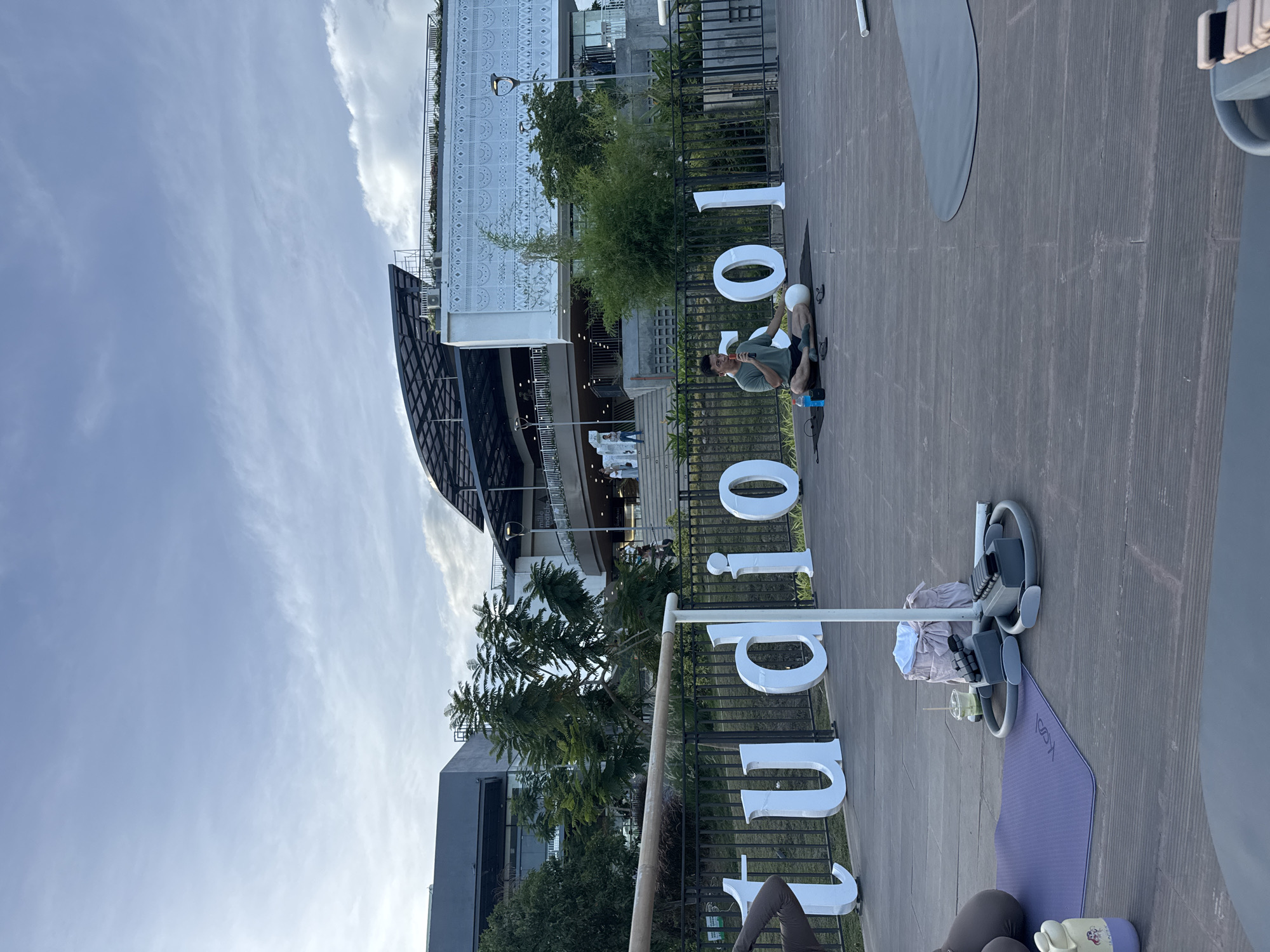 The image shows an outdoor space in Davao City, Philippines, with modern architecture and a large white sign spelling "lujo 101". A man wearing a green shirt and shorts sits on a black mat and appears to be speaking into a small red microphone. A small child sits next to him. Behind the man, the sign is situated in front of trees, buildings, and black metal fencing. To the right of the sign, exercise equipment (including pilates rings and a yoga mat) are on the ground. The sky is visible in the background and is partially cloudy.