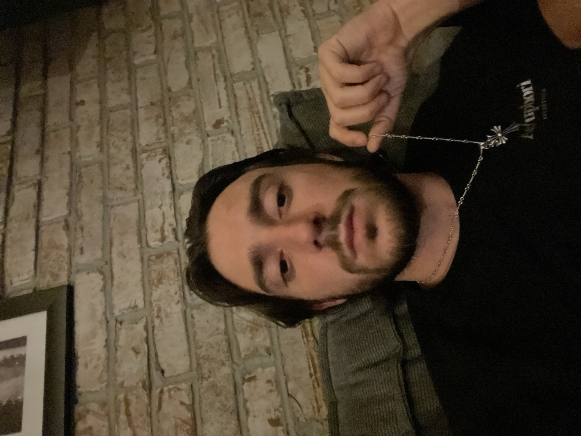 A man with dark hair and a beard is holding a silver necklace with a cross pendant in front of his chest. He is wearing a black t-shirt with white lettering. He is lying on a gray corduroy surface, likely a couch. Behind him is a brick wall. To the left of the wall is a framed picture. The location of the image in Huixquilucan de Degollado, Mexico is not discernable from the image.